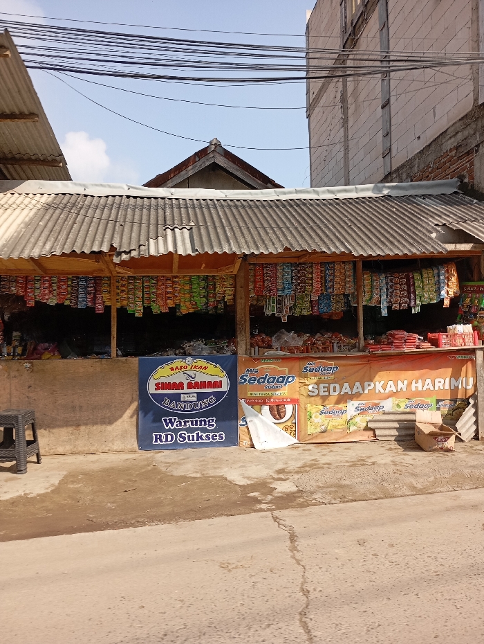 This image captures a street-level view of a small, open-fronted retail shop, commonly known as a "warung," located in Cipatik, Indonesia. The shop features a weathered, grey corrugated metal roof, supported by visible wooden beams. Numerous small, colorful packaged goods, including snacks and instant noodles, hang from lines beneath the roof, forming the primary merchandise display. Additional products are visible on shelves or surfaces within the shop interior.
Two promotional banners are affixed to the lower front portion of the shop. The banner on the left is blue, displaying white text for "BASO IKAN SINAR BAHARI BANDUNG" and "Warung RD Sukses." The banner on the right is orange, advertising "Mie Sedaaap" instant noodles with the slogan "SEDAAPKAN HARIMU."
The ground directly in front of the shop consists of a cracked concrete slab transitioning into packed dirt, showing signs of wear. A dark grey plastic stool is positioned to the left of the shop entrance. A flattened cardboard box rests near the shop's right side.
In the background, a multi-story, unpainted concrete building with visible vertical structural lines is present to the right, with a red brick wall at its base. A portion of another corrugated metal roof is visible to the far left. The sky overhead is clear blue with minimal cloud cover, and numerous black utility lines are visible crisscrossing the upper frame. No individuals are present, and no actions or interactions are depicted in the scene.