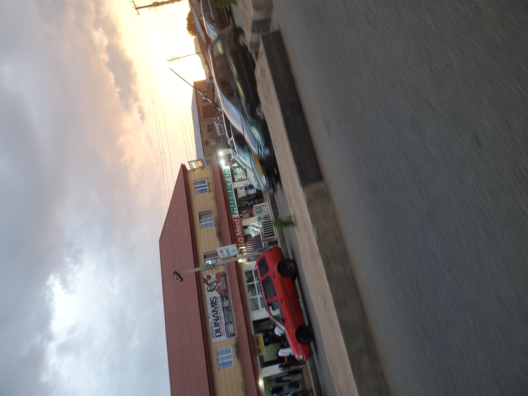 The image captures a street scene in Jos, Nigeria, during sunset. A two-story building with a red corrugated metal roof dominates the left side of the frame. The building features a sign that reads "WORSHIP OF DUNAMIS" with an image of two men, and another sign for "LA MED PHARMACY LTD." Several cars are parked and in motion on the road in front of the building, including a red station wagon, a dark green minivan, and a black sedan. A concrete barrier runs along the edge of the road, separating it from a sidewalk or another elevated area. Electrical poles and wires are visible, typical of urban infrastructure. The sky is filled with dramatic clouds, with the warm hues of sunset casting a glow on the horizon. The overall impression is one of a busy urban environment at the end of the day.