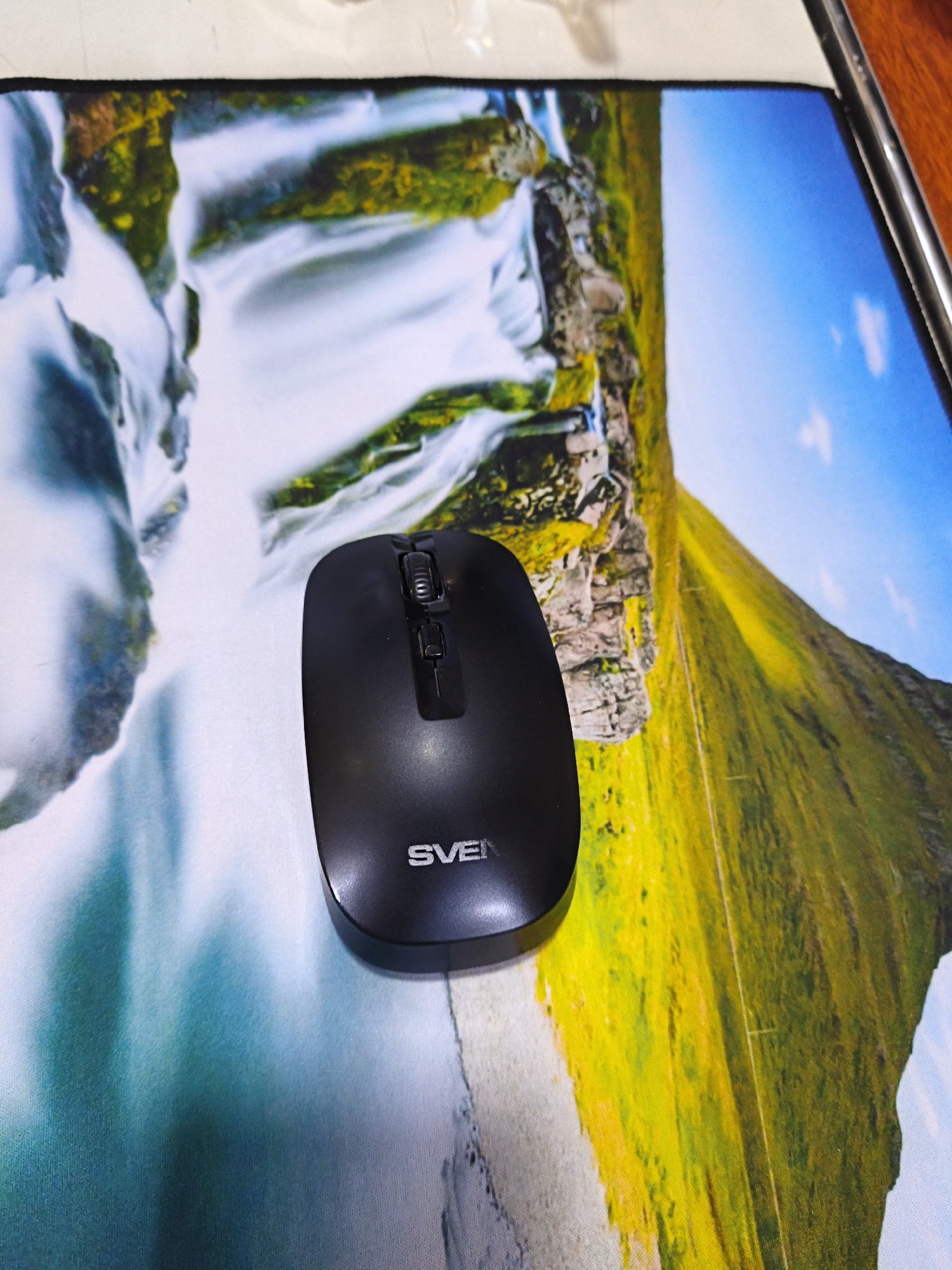 A black computer mouse is placed on a mousepad with a vibrant landscape scene featuring a waterfall and a grassy hillside under a blue sky. The mouse is positioned in the lower center of the image, with its scroll wheel and buttons clearly visible. The mousepad itself is colorful and detailed, creating a backdrop for the technology. The scene appears to be a close-up shot, with the mousepad covering most of the frame. There are no people or animals visible in the image. The location of Rishtan, Uzbekistan is not depicted in this image.