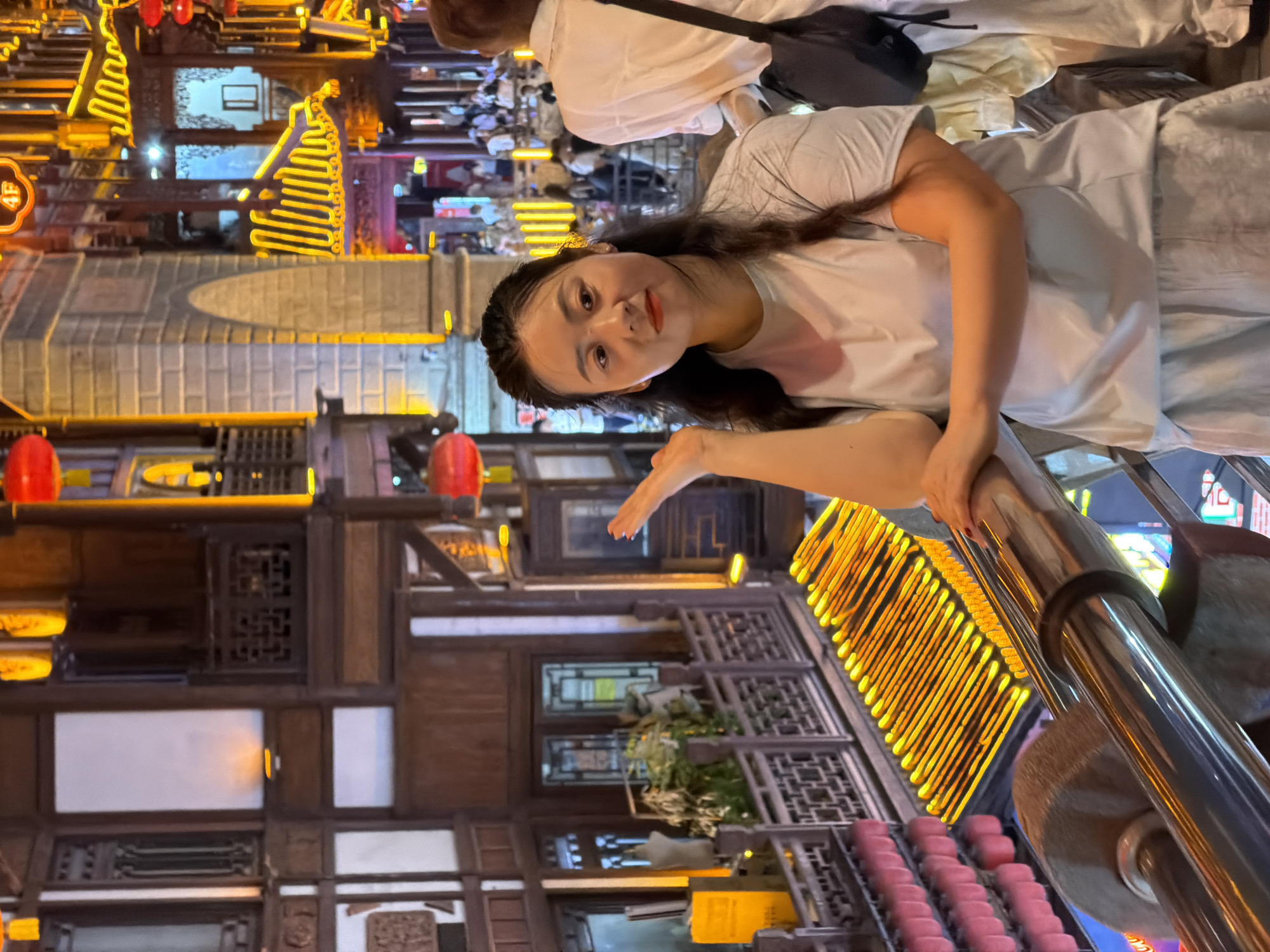 A medium shot captures a young Asian woman standing on a balcony overlooking a busy street at night, likely in Yuzhong District, China, given the traditional architecture and illuminated lanterns. The woman, with dark hair pulled back and wearing a light blue t-shirt and light-colored shorts, is looking towards the camera with a slight smile. Her right arm is extended, hand open, as if gesturing. Behind her, a man in a white shirt and dark pants stands with his back to the camera. The background features multi-story buildings with ornate wooden balconies, traditional Chinese architectural elements, and vibrant yellow and red lights, suggesting a tourist area or entertainment district. The street below is filled with people, and the scene is illuminated by the warm glow of numerous lanterns and neon signs.