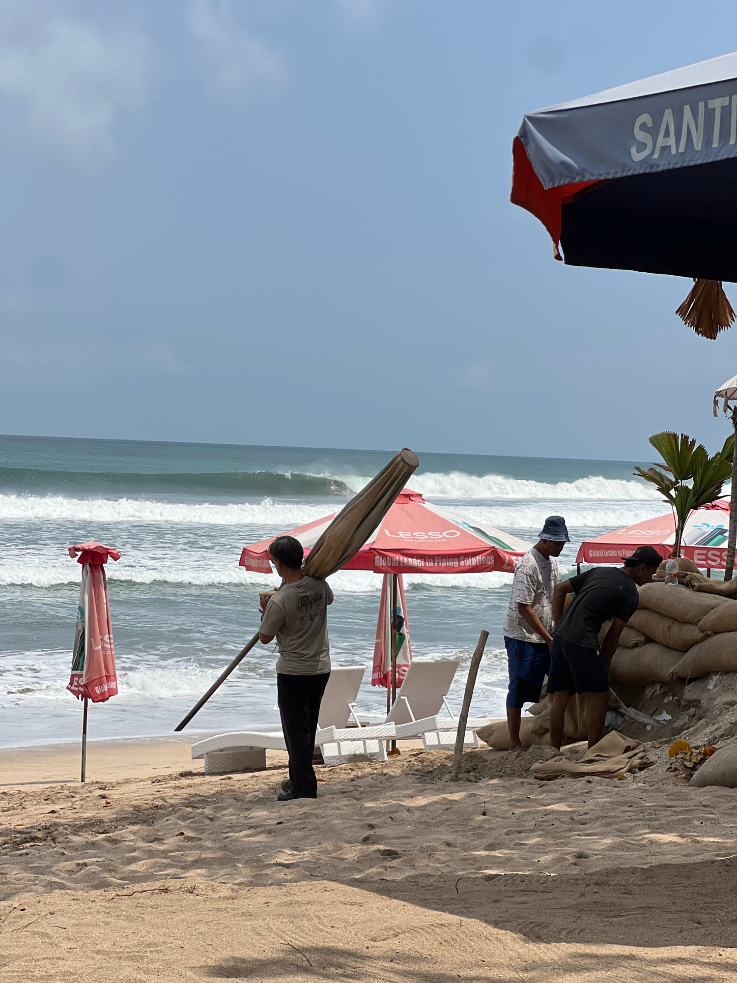 On a beach in Kuta, Indonesia, several people are working near the water. In the foreground, a man carries a large, rolled-up umbrella over his shoulder. Further back, two men are interacting near a wall of sandbags. One man is wearing a black t-shirt and dark shorts, and appears to be kneeling, possibly working on the sandbags. The other man, in a patterned t-shirt and blue shorts, stands nearby. The ocean has moderate waves, and several beach umbrellas are visible. One umbrella in the background has the word "SANTI" printed on it. The sky is a pale blue with some clouds. The beach is sandy.