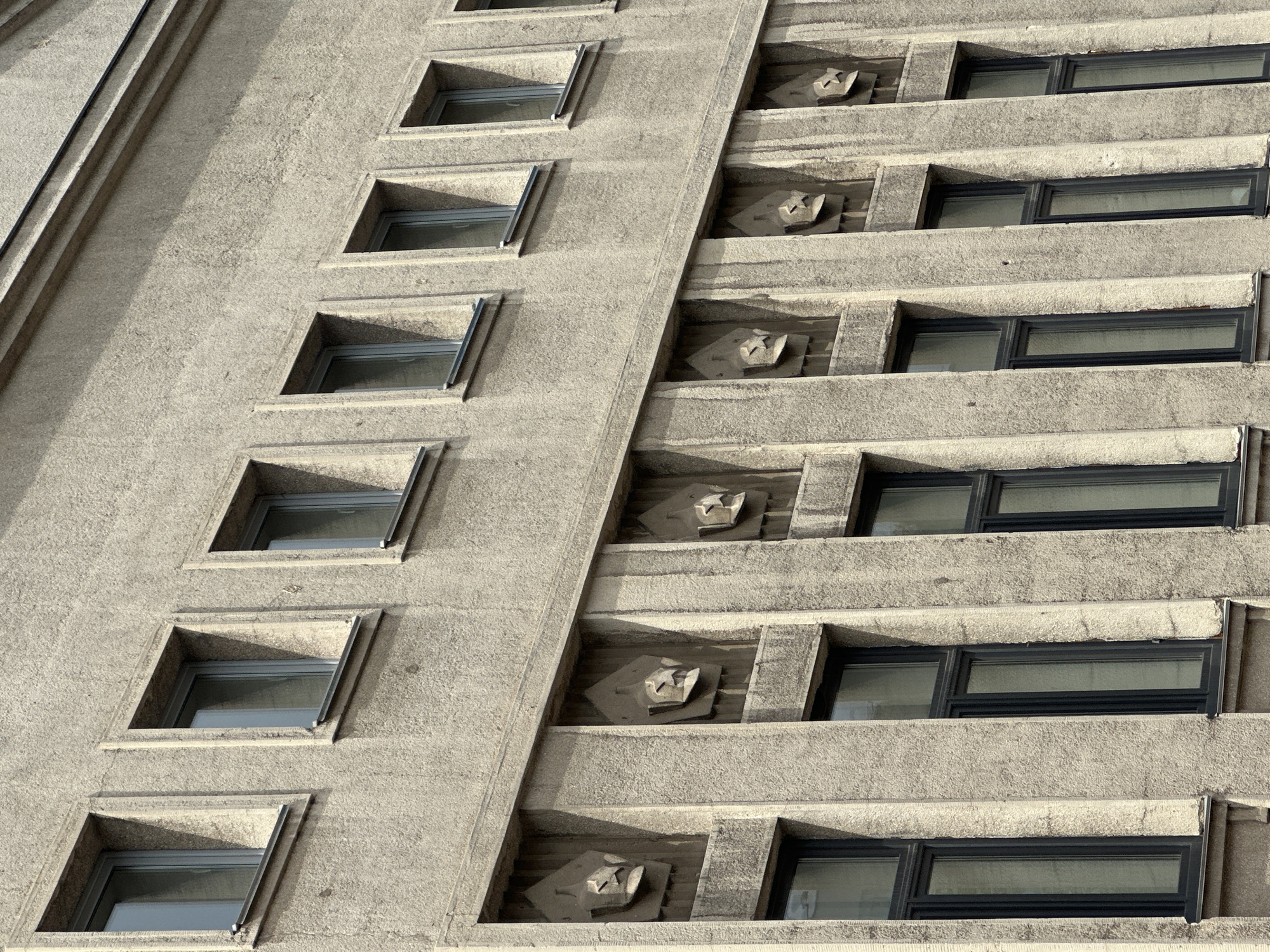 The image shows a section of a Brutalist architectural building in Minsk, Belarus. The building's facade is constructed of rough concrete with a textured finish. A series of uniformly spaced rectangular windows are visible across two horizontal levels. The upper level windows are smaller and wider, while the lower level windows are taller and narrower. Between the two levels of windows, a decorative frieze runs horizontally across the facade. This frieze is composed of repeating relief sculptures, each depicting a stylized Soviet-era military helmet or cap with a star emblem. The overall impression is one of stern, monumental design characteristic of Soviet public architecture. No people or signs of activity are visible in the frame.