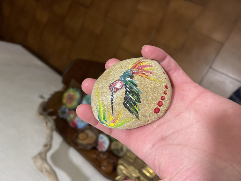 A close-up shot features a person's hand holding a smooth, light-colored, oval-shaped rock, which has been artfully painted. The central subject of the painting is a vibrant bird, possibly a stylized hummingbird or a mythical creature. It has a slender black beak, a white and red head, and a body adorned with shades of green and black. A striking crest of red and yellow strokes radiates from its head, and green and yellow brushstrokes at the base suggest foliage. Along the right edge of the rock, a series of small, bright red dots forms an arc.
The hand gently cradles the painted stone, presenting the artwork directly to the viewer. In the softly blurred background, several other painted rocks or small decorative objects are visible within what appears to be a wooden tray or shallow container. The setting seems to be indoors, under warm artificial lighting, with the background suggesting a domestic space featuring brown-toned surfaces, possibly wood or tiles. The image was captured in Dosso (Ciappanico), Italy, showcasing a piece of handcrafted art. No visible text is present in the frame.