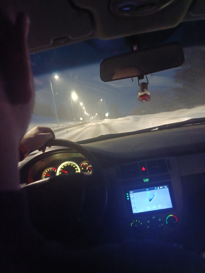The image captures a first-person perspective from inside a car, looking out through the windshield onto a snowy road at night. The setting suggests a winter evening drive.
Inside the car, a person's left arm and hand are visible in the foreground, holding the steering wheel, indicating that the viewer is in the driver's seat of a left-hand drive vehicle. The dashboard is illuminated with warm orange and yellow lights for the gauges, including a speedometer, tachometer, and fuel/temperature indicators. The speedometer needle points to approximately 60-70 units, likely kilometers per hour.
Below the dashboard, on the center console, a red triangular hazard light button is visible. A digital clock displays "7:09". Dominating the center console is a modern, blue-lit touchscreen infotainment system, appearing to show a media playback interface with a graphical waveform or sound representation. Below the screen are controls for the car's climate system, also illuminated with a green glow. Hanging from the rearview mirror is a distinctive car air freshener or ornament, featuring a red, multi-faceted base and a golden or bronze, intricately shaped top, possibly resembling a crown. The rearview mirror itself is dark.
Outside the car, the road ahead is covered in snow, with visible tire tracks. Numerous bright streetlights line both sides of the road, receding into the hazy distance, creating a sense of depth and illuminating the snowy landscape. The bright lights suggest a significant road or highway. The ground on either side of the road is also covered in snow, and a guardrail is visible along the left side. The overall atmosphere is cold and wintry, illuminated by artificial light sources. The faint haze in the air might indicate falling snow or fog.
