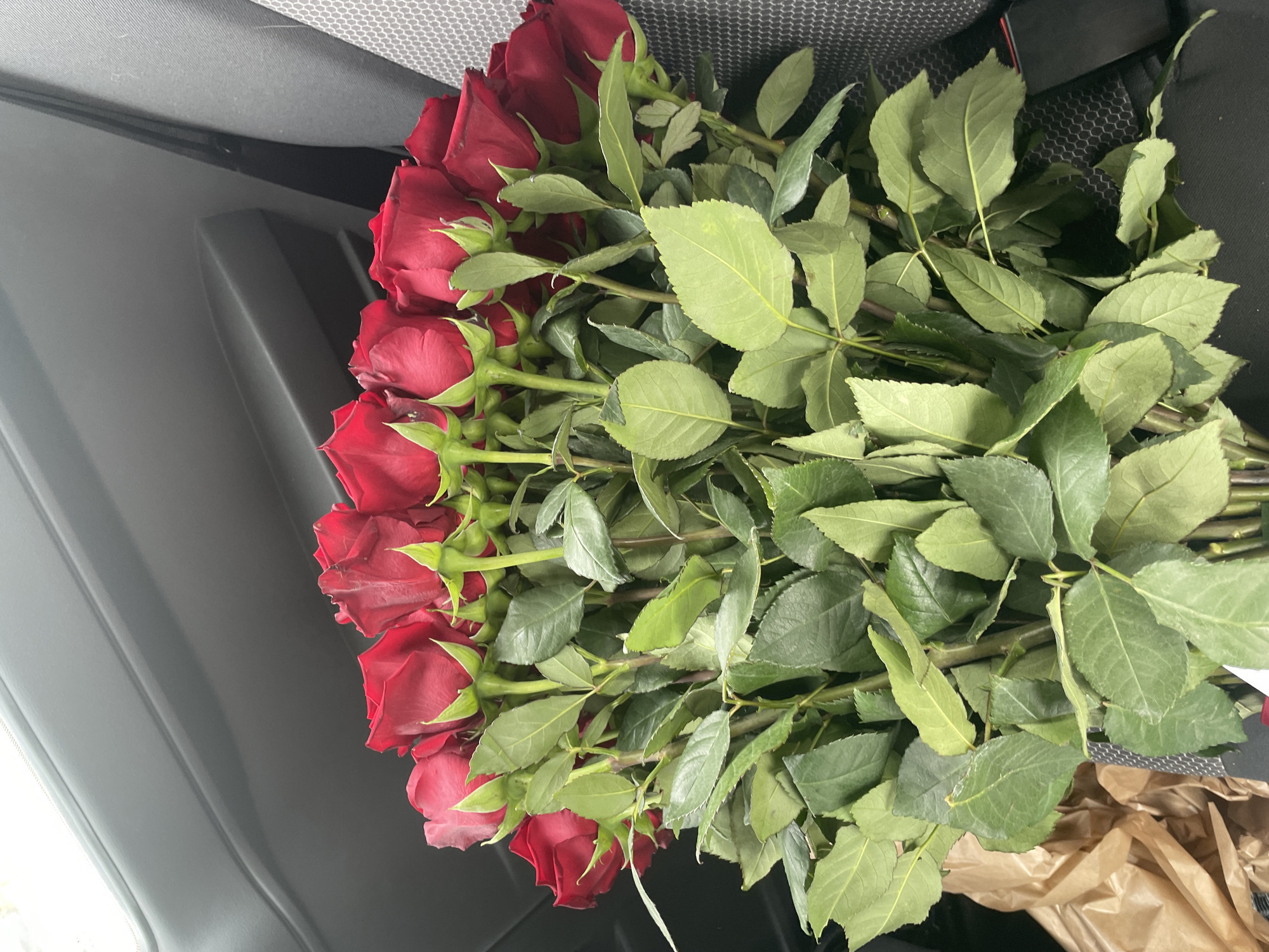 The image shows a bouquet of red roses lying on a dark surface inside a vehicle. The roses are clustered together with their green leaves and stems visible. The flowers appear fresh and vibrant, with the deep red color of the petals contrasting against the green foliage. The background reveals parts of the car interior, including a dark dashboard or console and possibly the seat or lining. The scene suggests the flowers are being transported, possibly as a gift or purchase.