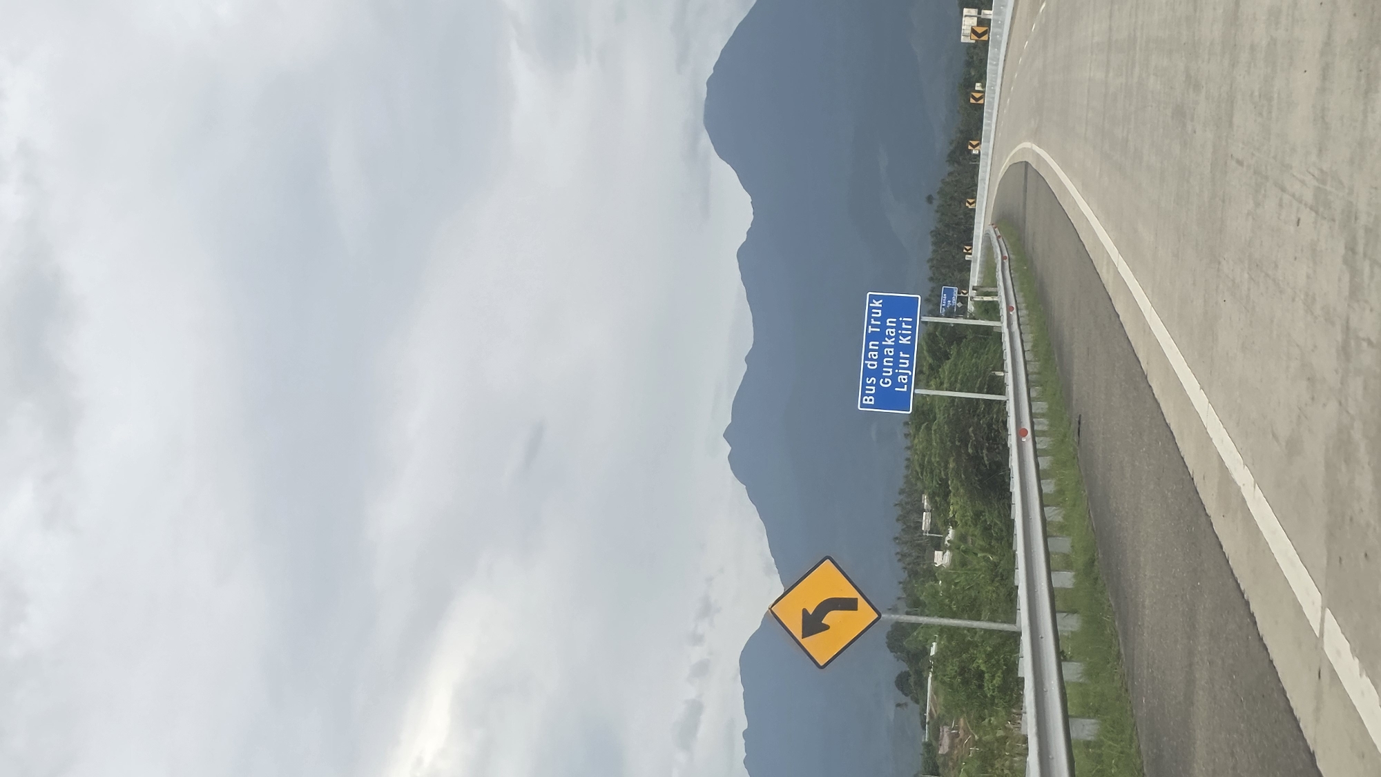 The image shows a highway in Baksukon, Indonesia. A gray asphalt road curves to the right, with a white lane marker dividing it. A gray metal guardrail runs along the edge of the road. On the left side of the road, two road signs are visible. One is yellow and diamond-shaped, with a black arrow curving to the left. The other is blue with white text in Indonesian, which translates to "Buses and Trucks Use Left Lane." In the background, mountains are visible under a cloudy sky. Green vegetation is seen between the road and the mountains.