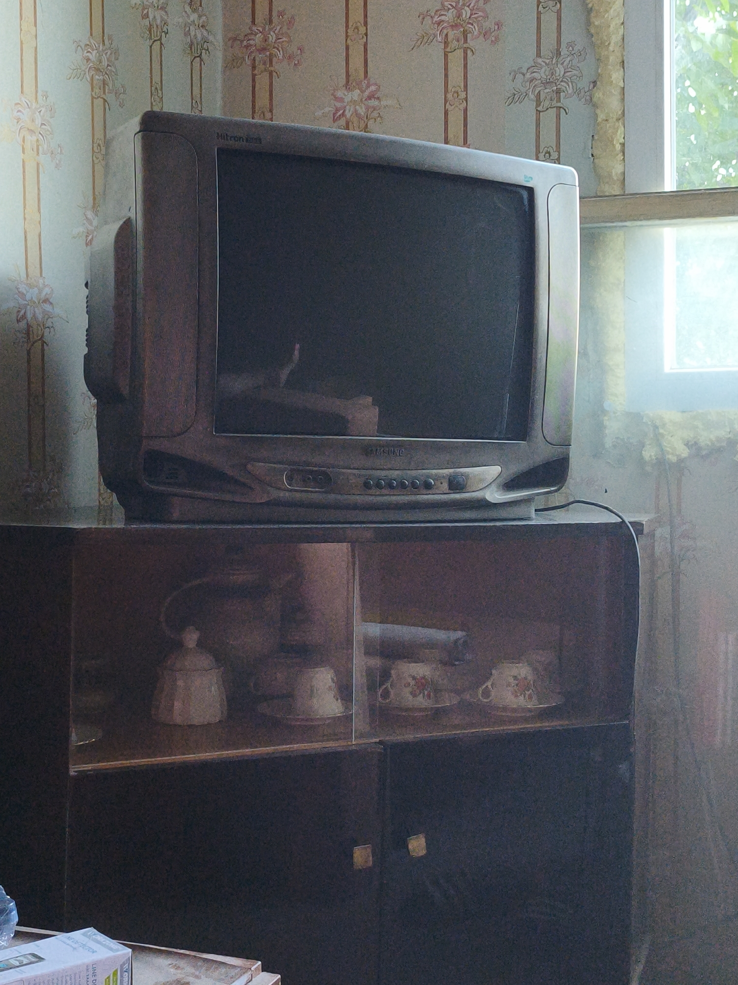 The image depicts an interior scene, likely a living room, within a dwelling in Lovech, Bulgaria. In the foreground, a dark wooden cabinet with glass-fronted shelving is visible. Inside the cabinet, a white lidded jar or sugar bowl and several teacups with floral patterns are arranged. The cabinet appears to be made of particleboard or a similar material, with two small square knobs on the lower doors.
On top of the cabinet rests an older model television set. The television is gray and has a prominent bezel. Text above the screen appears to read "Hitron" and possibly "Gold". Below the screen, there are control buttons and a slot. A black cable emerges from the right side of the television and curves downwards.
The background features wallpaper with a floral pattern of pink and cream flowers against a light green and yellow striped background. To the right of the television, a window is visible, showing a blurred view of green foliage, suggesting an outdoor environment. The lighting in the scene is somewhat dim, with light entering from the window. The overall impression is of a lived-in, perhaps slightly dated, domestic space.