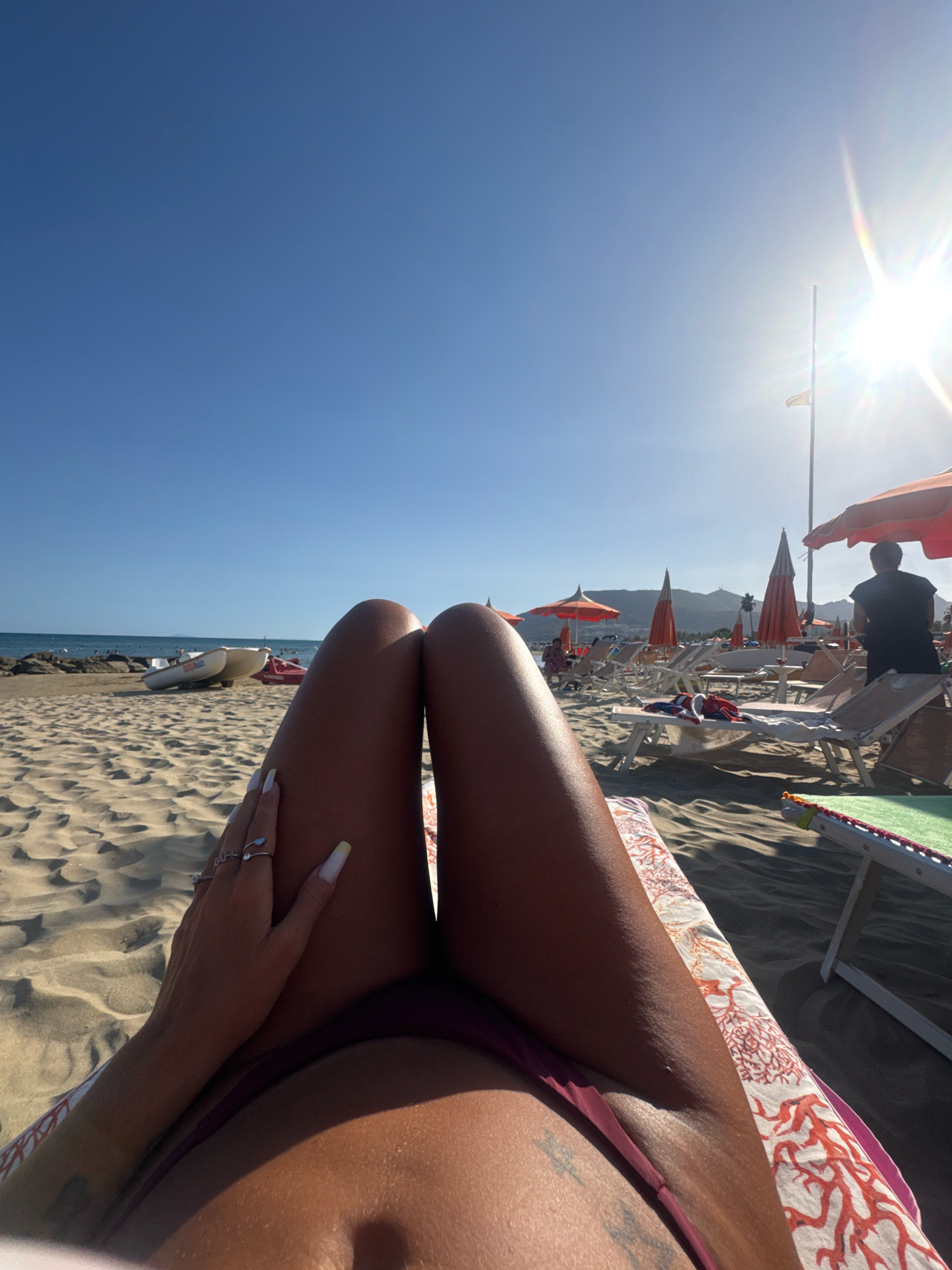 The image depicts a point-of-view shot of a person lying on a beach lounger on a sunny day. The foreground focuses on the person's tanned legs and hand, adorned with rings and long, white-tipped nails. The person is wearing a dark-colored bikini bottom.
The background reveals a sandy beach with rows of empty lounge chairs and orange umbrellas. In the distance, the sea meets a clear blue sky, with mountains visible on the horizon. A person in a dark shirt is standing near some of the umbrellas, facing away from the camera. Two small white boats are resting on the sand near the water's edge. The scene is illuminated by bright sunlight, creating lens flare in the upper right quadrant of the image. The location is identified as San Felice Circeo, Italy, suggesting this is a beach resort area.