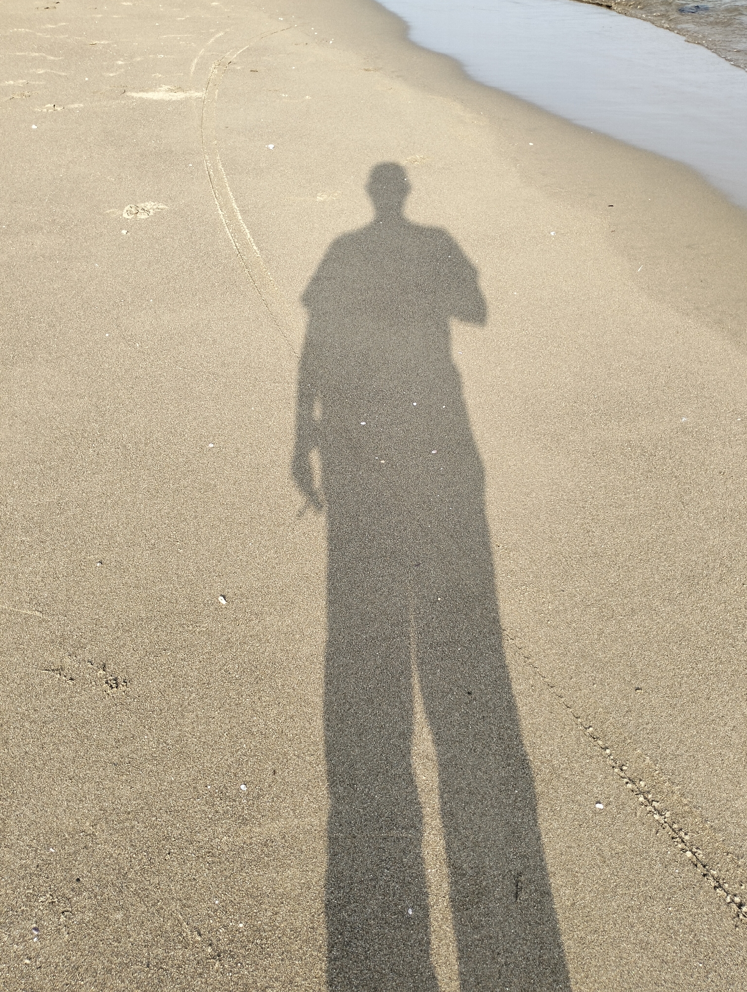 A shadow of a person is cast upon a sandy beach in Curehtunong, Indonesia. The sand is predominantly tan, transitioning to a darker hue near the water's edge. The ocean is visible in the upper right of the frame. There are shells and beach debris scattered around.
