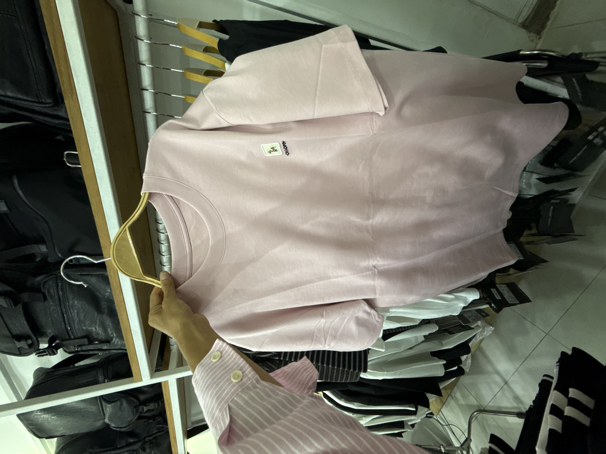 A person's hand, wearing a pink and white striped long-sleeved shirt, holds up a light pink t-shirt on a wooden hanger. The t-shirt is plain with a small embroidered logo on the left chest that reads "CHAOPIN". The t-shirt is on display in a retail setting, with other clothing items and black backpacks visible in the background. The scene appears to be in a store or market in Nha Trang Ward, Vietnam.