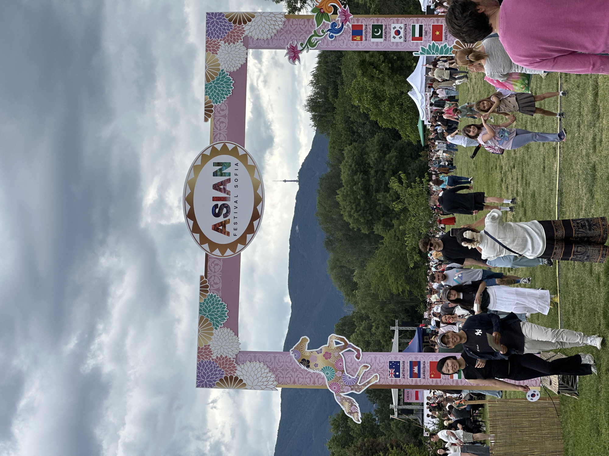 This is an outdoor photograph of the Asian Festival in Sofia, Bulgaria. A large, decorative archway, adorned with floral motifs and a stylized horse, stands at the entrance to the festival grounds. The archway features the text "ASIAN FESTIVAL SOFIA". The background shows a lush green park with trees, mountains, and a distant tower. Numerous people of various ages are walking through the archway and milling about the festival. There are also flags representing different countries displayed vertically on the right side of the archway. The sky is overcast with dramatic clouds. The overall scene depicts a lively public event.