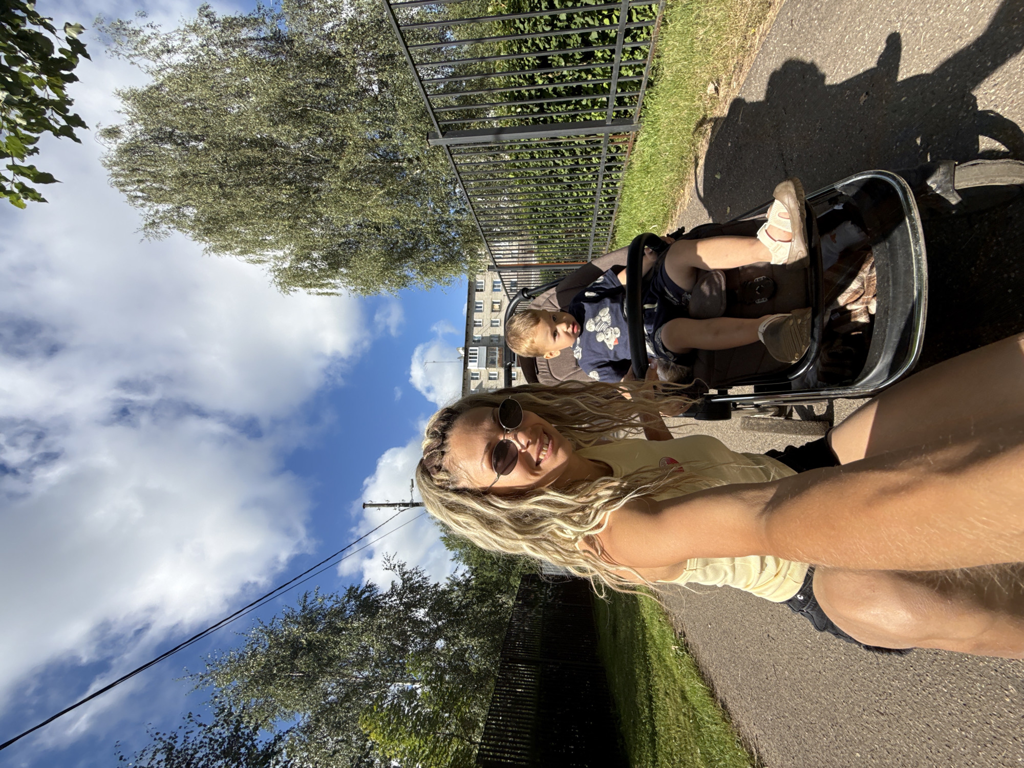 An outdoor, low-angle, eye-level, wide shot captures a woman and a young boy in a stroller on a paved path under a partly cloudy sky. The woman, in the foreground, has blonde dreadlocks, is wearing sunglasses and a yellow tank top with black shorts, and is smiling at the camera. The young boy, seated in the stroller, wears a blue shirt with a graphic print and dark shorts. The stroller is brown with a silver frame. Behind them, a black metal fence runs along a grassy area, with trees and a multi-story building visible in the distance. The scene appears to be in Rusanovo, Russia, indicated by the presence of power lines and a typical Russian apartment building. The lighting suggests it is daytime, possibly late morning or early afternoon, with the sun casting shadows.