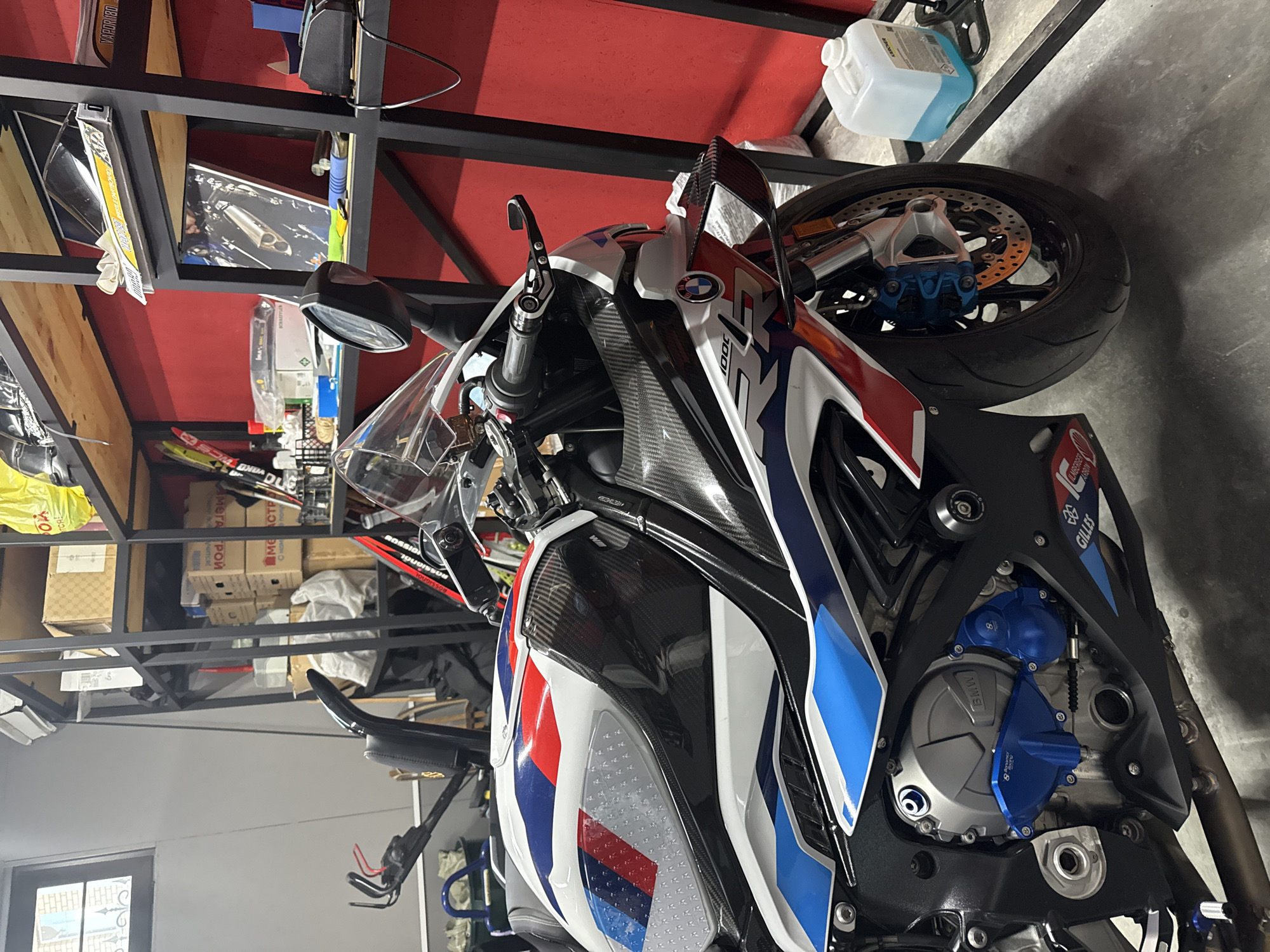 The image shows a BMW S 1000 RR motorcycle in a garage or workshop setting. The motorcycle is characterized by its white, red, and blue livery, carbon fiber accents, and high-performance components such as blue-colored engine covers and brake calipers. The background consists of shelving units containing various items, including boxes and containers. The location is likely a private garage or workshop, and there's no information to specify the city of Дубровка, Russia.