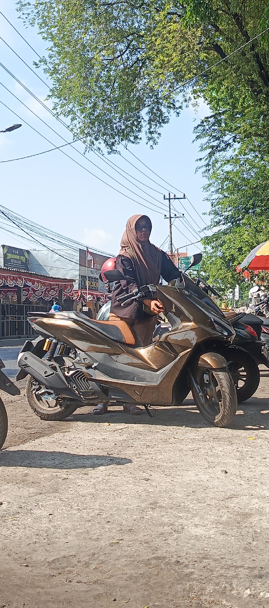 In Krian, Indonesia, a woman wearing a brown hijab and sunglasses is seated on a bronze-colored scooter on a sunny day. She is wearing a dark long-sleeved top and brown pants. The scooter has a red helmet resting on the handlebars. The background shows trees, utility poles with numerous wires, and buildings along a street. One building has signs that are partially visible, including one that reads "COMMANDER WISH." Another sign is partially obscured by a red and yellow umbrella. The ground is a rough, light-colored surface, likely asphalt or concrete, with shadows cast on it. Other parked motorcycles are visible to the right of the main scooter.