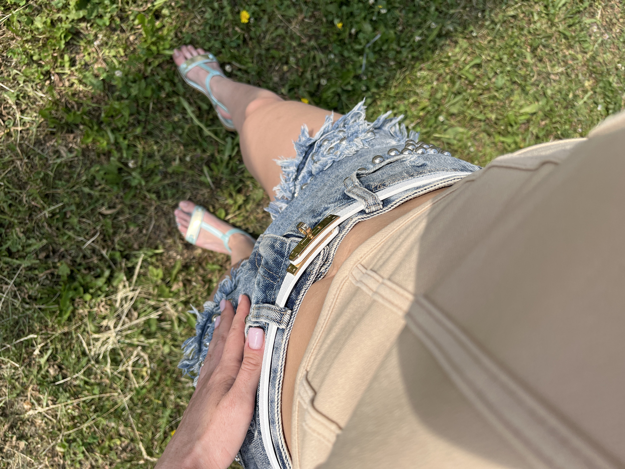 The image is a close-up, overhead view of a person's lower body, seated on a grassy area. The person is wearing a beige garment on their upper legs and a distressed denim skirt with a white belt and gold buckle. Their feet are visible, wearing light blue and white sandals. The background consists of green grass with scattered weeds and yellow flowers, suggesting an outdoor setting in a park or garden. The lighting indicates it is daytime, with shadows cast by the sun. The specific location within the city of Kazan, Russia, cannot be determined from the visual information provided.