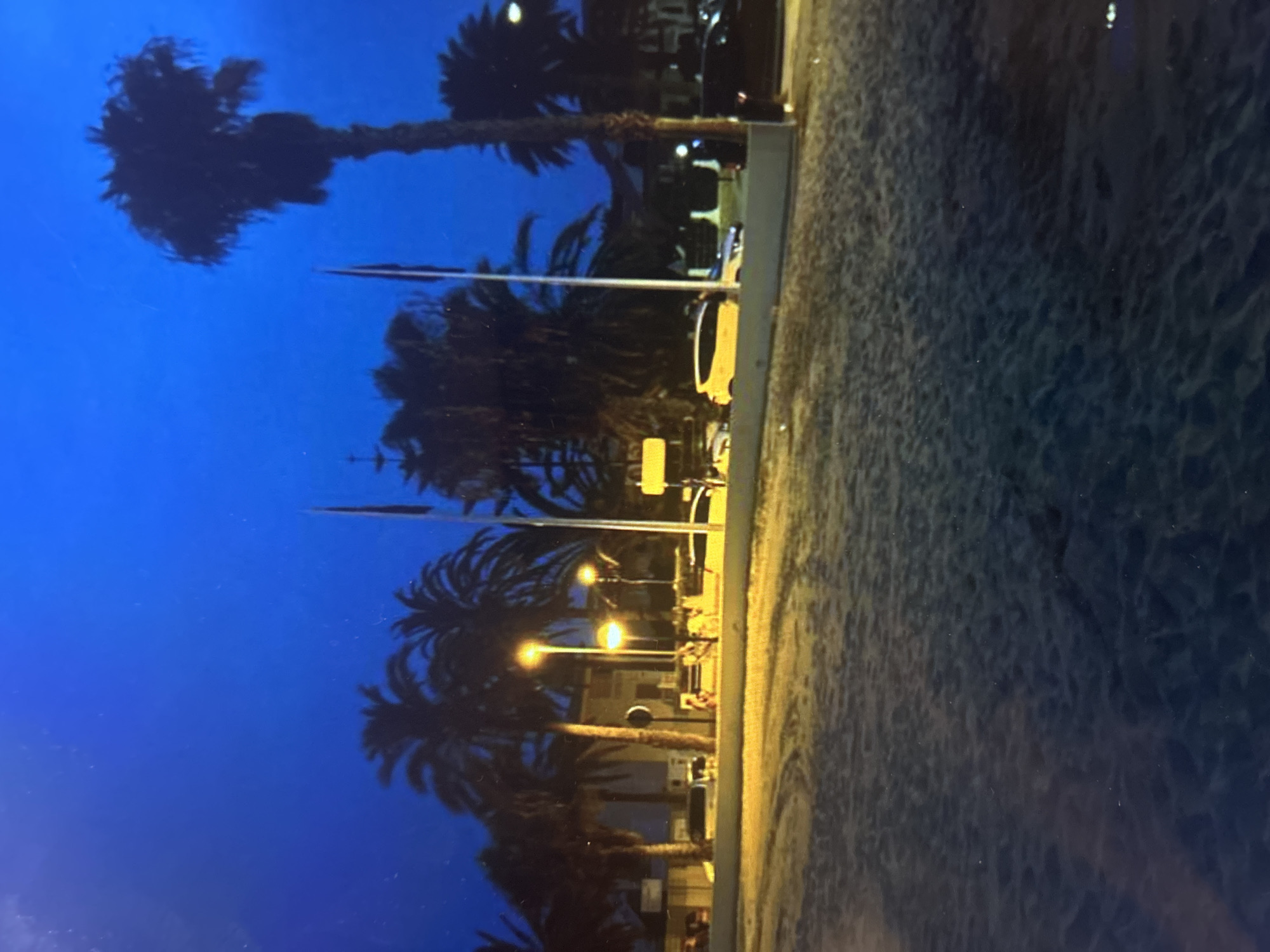 The image depicts an outdoor night scene in a coastal urban environment. The foreground consists of textured sand, likely a beach, extending across the bottom half of the frame. A low, light-colored concrete barrier separates the sand from a paved area or road in the middle ground. Along this paved area, several illuminated streetlights with warm yellow light are present. Numerous tall palm trees, silhouetted against the dark blue sky, dominate the middle ground and background.
Behind the barrier, on the paved surface, multiple vehicles are parked. A bright yellow hatchback car is prominently visible on the right, facing left. To its left, a lighter-colored car is partially visible. Further right, a dark-colored car is partially obscured by a palm tree trunk. In the background, multi-story buildings with visible windows are situated amongst more palm trees. Three tall poles are visible in the middle ground, two bearing dark, rectangular flags or banners. The sky is a consistent dark blue, characteristic of twilight or night. No individuals or specific activities are discernible.