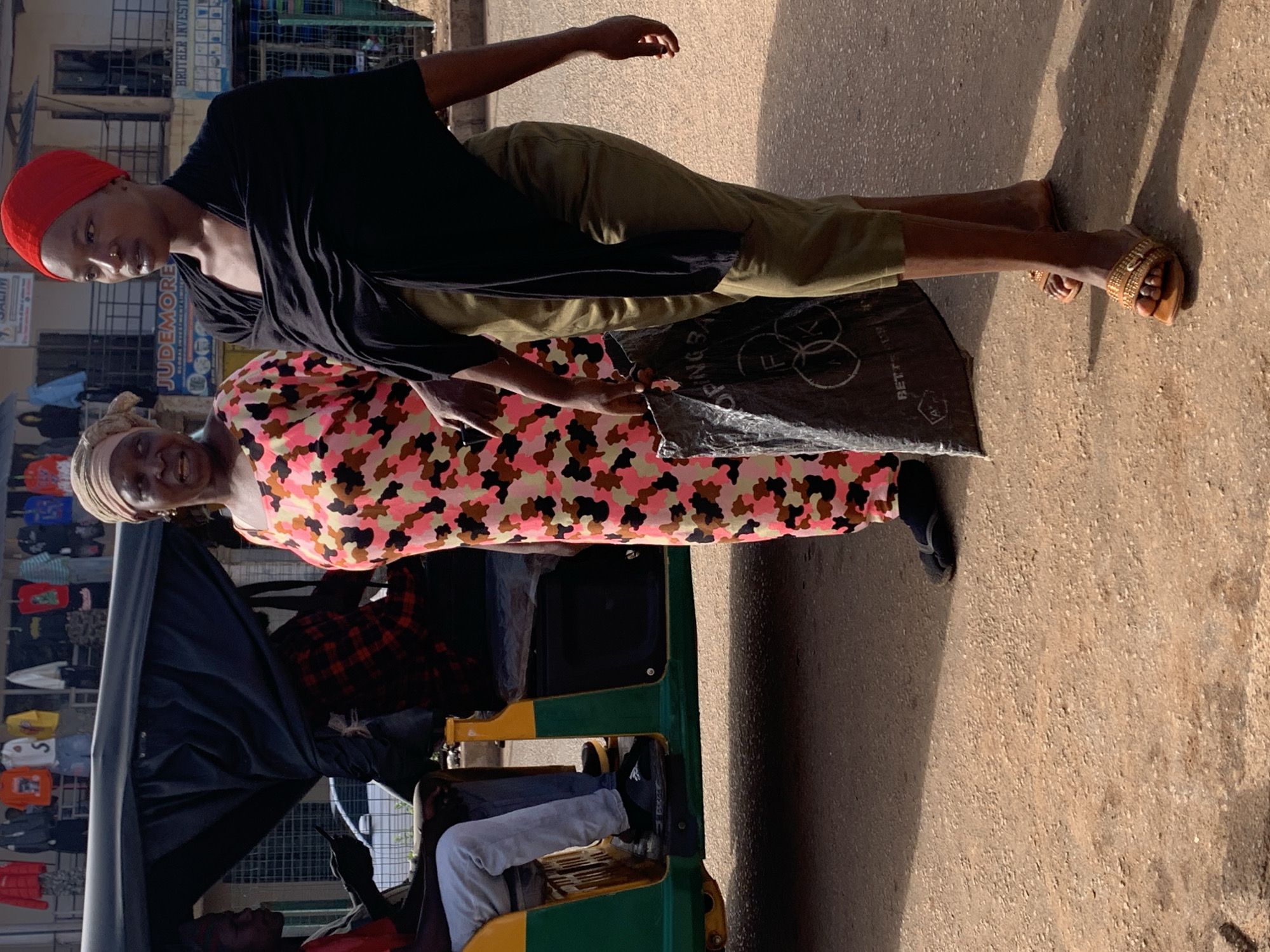 The photograph captures a street scene in Bukuru, Nigeria. Two women are the central subjects. The woman on the left, facing the camera with a slight smile, wears a headscarf and a colorful, camouflage-patterned dress. To her right, a younger woman with a red headscarf and dark clothing is looking away from the camera. She is carrying a large black plastic bag. In the background, a three-wheeled vehicle, possibly a motorized rickshaw, is visible, with a person seated inside. The setting appears to be a marketplace or busy street, with shops and awnings lining the background and merchandise displayed on racks. The ground is dusty and uneven, typical of unpaved urban areas. The lighting suggests it is daytime, with harsh shadows indicating sunlight.