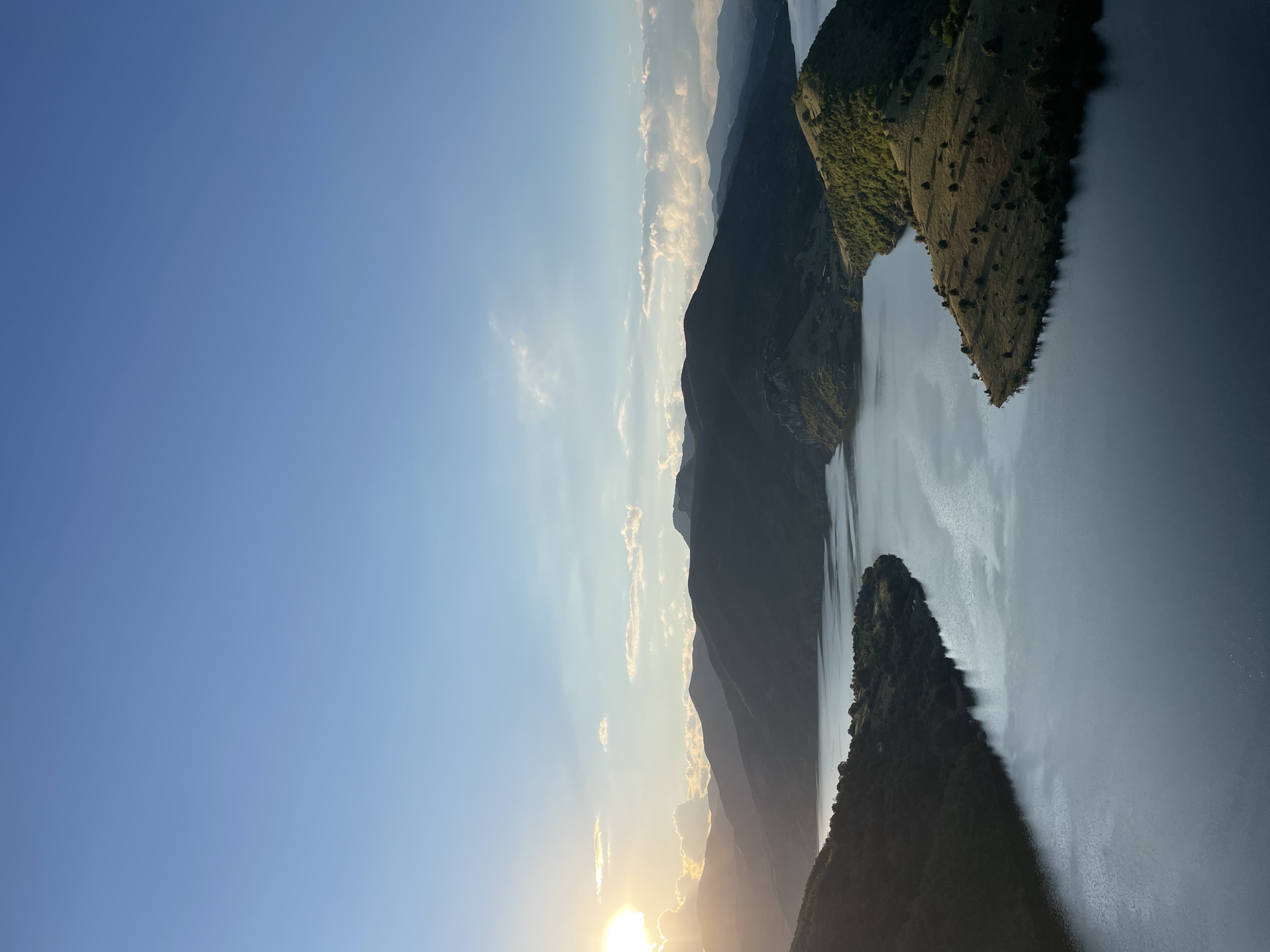 The image captures a scenic view of a lake and surrounding mountains. The sun is setting on the left side of the frame, casting a warm glow over the scene and creating a silhouette of the mountains. The lake's surface reflects the sky and the surrounding landscape, with ripples indicating gentle movement of the water. The foreground shows a peninsula jutting out into the lake, covered with dry grass and scattered trees, with a dense green forest on the opposite side. The background is dominated by layers of mountains receding into the distance, some covered with trees and others appearing more barren. The sky is a clear blue with a few scattered white clouds, tinged with orange and yellow from the setting sun. The location is identified as near Ribartsi, Bulgaria.