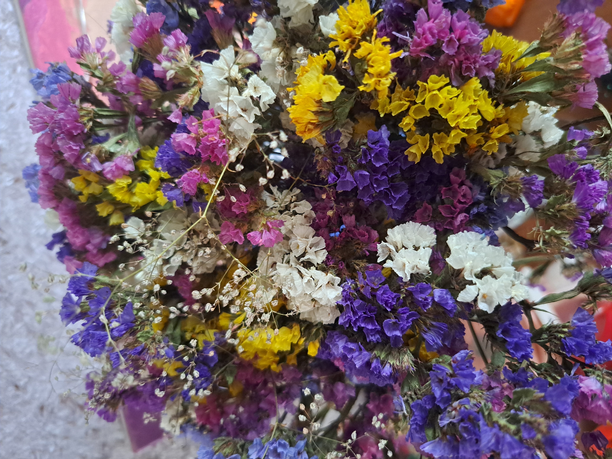 A close-up, overhead shot shows a bouquet of dried flowers in Vypasne, Ukraine. The flowers are a mix of colors including purple, blue, yellow, white, and pink. They are arranged in a dense cluster, with some stems and greenery visible. The background is blurred, but appears to be a textured, light-colored surface.