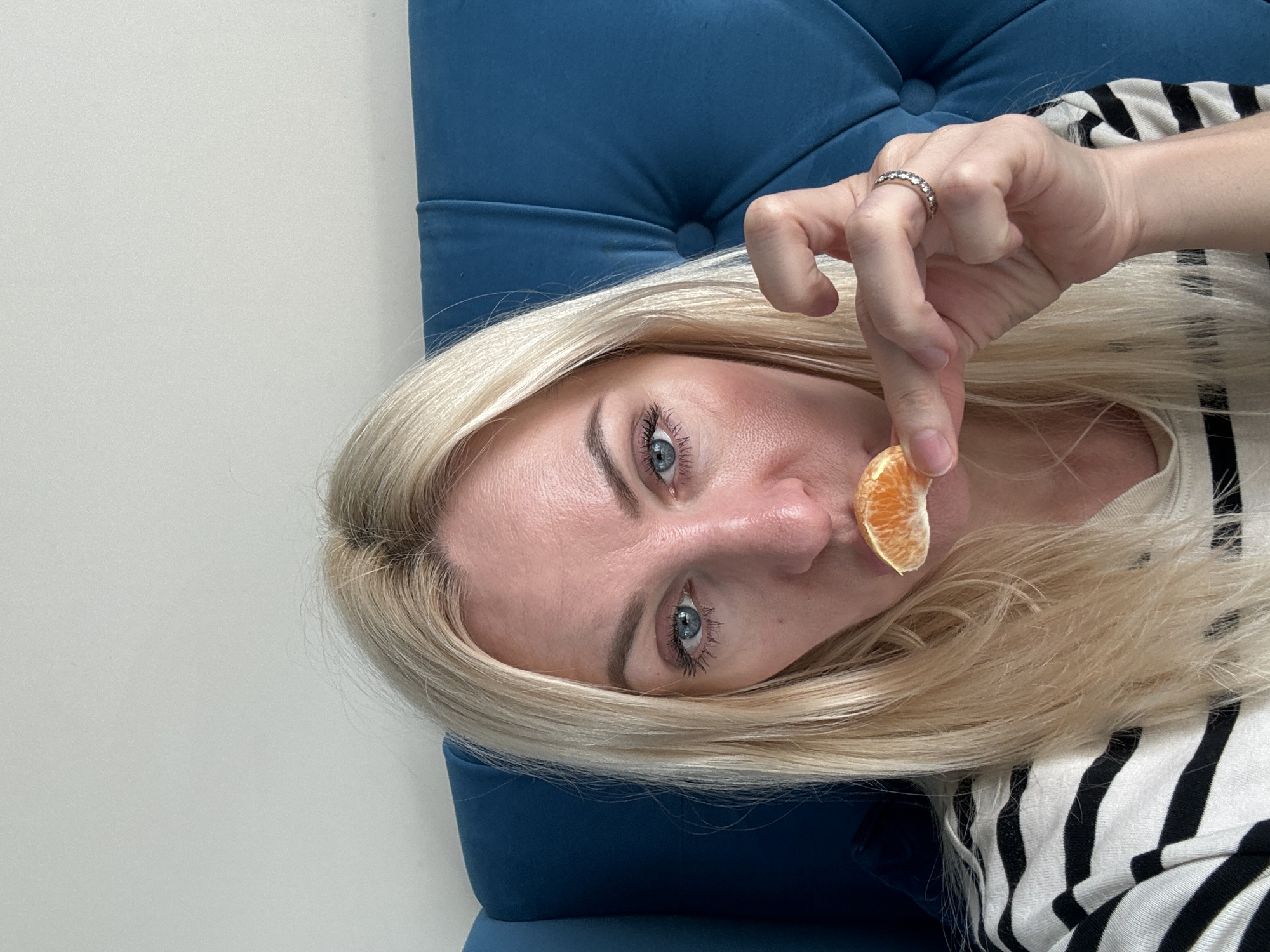 A close-up, waist-up shot captures a woman with blonde hair and blue eyes, holding a segment of an orange up to her lips. She is wearing a black and white striped shirt and is seated against a textured blue headboard. The background is a plain white wall. The overall impression is of a casual, indoor portrait.