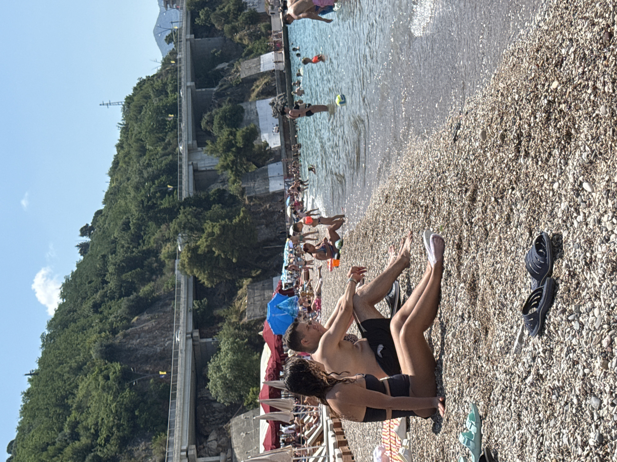 The image shows a crowded pebble beach on a sunny day, likely in Boreti, Montenegro. The water is calm and clear, with several people swimming and wading. On the beach, many people are sunbathing, sitting, and walking. There are beach umbrellas providing shade, and lounge chairs scattered around. In the background, a steep, tree-covered hillside rises, with a modern-looking elevated road or walkway running along its edge. A few yellow flags are visible along the elevated structure. The overall atmosphere appears to be relaxed and leisurely, typical of a summer beach scene. The time of day is likely midday or early afternoon due to the bright sunlight and strong shadows. No visible text is present in the image.