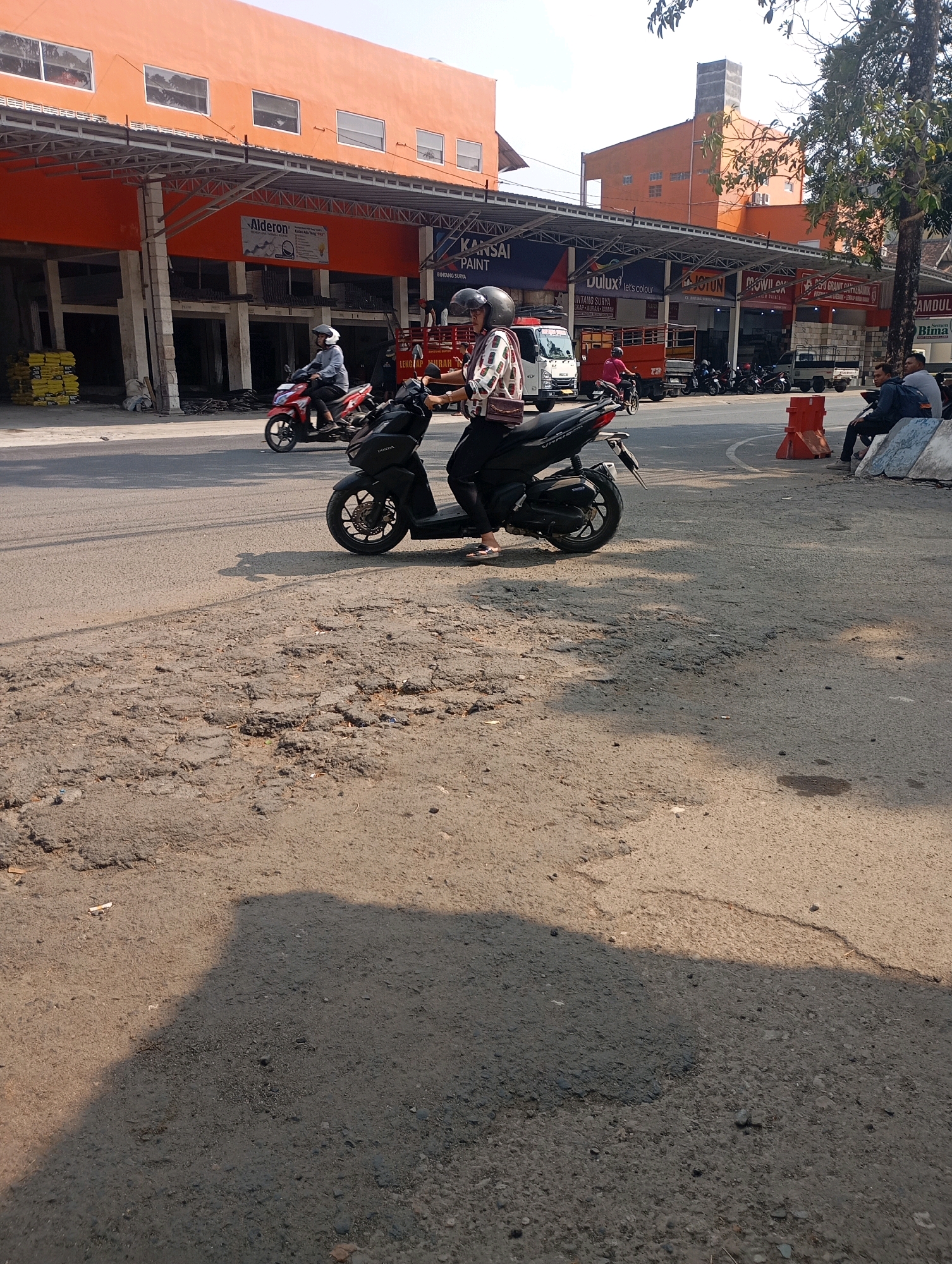 The image depicts a street scene in Pacitan, Indonesia, with a focus on a woman riding a black Honda scooter. The scooter is in the foreground, partially obscuring a damaged asphalt road with visible potholes and cracks. The woman is wearing a white and brown patterned shirt, black pants, and a black helmet. Her feet are visible, wearing flip-flops. She is riding towards the right of the frame.
In the background, an orange building with white window frames lines the street. Several signs are visible on the building's facade, including one for "Alderon" and another for "Kansai Paint." Several other vehicles are present on the road, including a red scooter to the left of the main subject, a white truck, and several parked motorcycles. On the right side of the road, a group of three men are seated on what appears to be a low wall, some of them wearing casual clothing. Orange traffic cones are also visible on the right side of the road, near the men. The overall environment suggests a commercial area on a sunny day, with visible tree foliage in the upper right portion of the image.