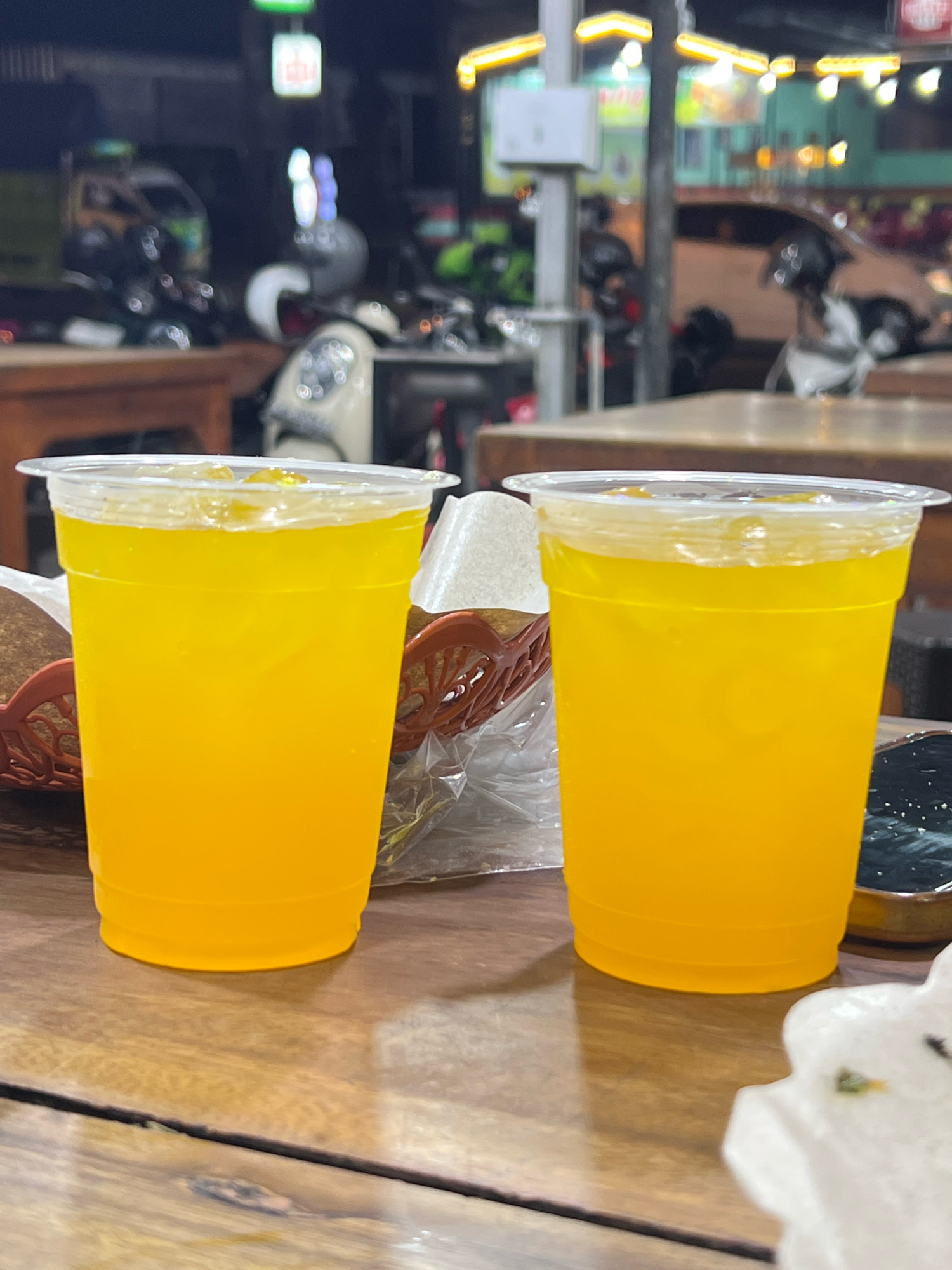 The image shows two plastic cups filled with a bright orange beverage, likely juice or iced tea, sitting on a wooden table. The cups are clear and have white plastic lids. In the background, the scene appears to be an outdoor dining area at night, possibly in Denpasar, Indonesia, given the presence of neon signs and what looks like a street scene with motorcycles and a vehicle. The lighting is dim, with some artificial light sources casting a glow. One of the cups is closer to the foreground and more in focus, revealing ice cubes within the drink. To the left of the cups, a brown woven basket is partially visible. To the right, a dark, possibly ceramic, dish is partially visible. The overall impression is of a casual evening setting, likely at a restaurant or street food stall.