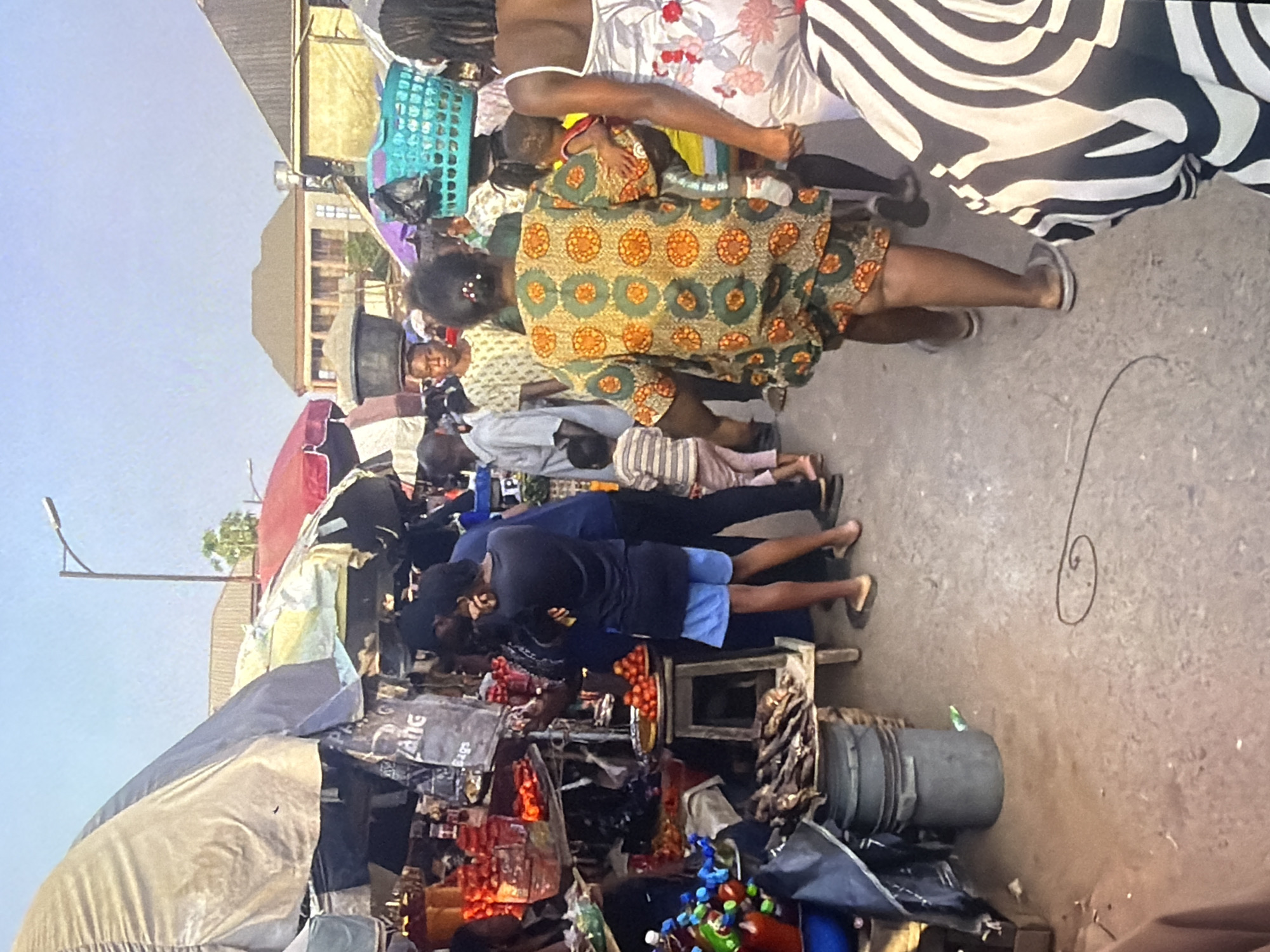 The image depicts a bustling outdoor market scene in Bukuru, Nigeria. The foreground is dominated by the lower bodies of several people, including one individual wearing a black and white striped garment and another with legs adorned in a patterned green and orange fabric. Various market stalls with awnings are visible, displaying goods such as tomatoes, dried fish, and bottled beverages. Several individuals are walking through the market, some carrying items, including a woman with a blue basket on her head and a child. Buildings with tiled roofs and a street light are discernible in the background. The ground is paved with concrete. The overall impression is one of active commerce and daily life in a Nigerian urban environment.