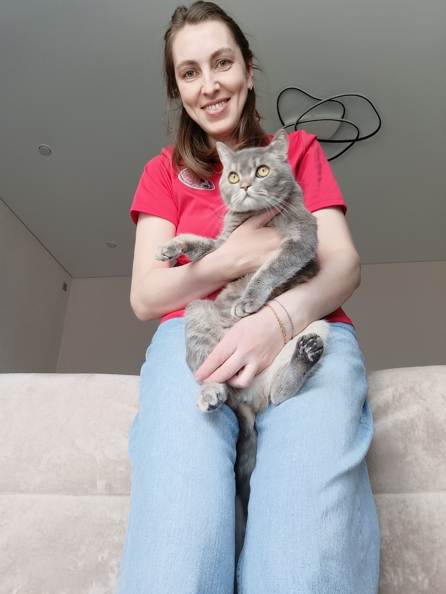 The image captures a smiling woman holding a grey tabby cat in an indoor setting, likely in Chaikovsky, Russia. The woman, appearing to be in her late 20s or 30s with light brown hair pulled back, is seated on a light-colored sofa. She wears a red short-sleeved t-shirt and light blue jeans. On the right side of her t-shirt, a circular emblem is visible, featuring partially discernible text such as "WORLD CUP" and a design that includes stars and stripes, resembling a flag. She also wears two delicate chain bracelets on her left wrist.
The grey tabby cat, with prominent yellow eyes, is nestled in her arms, looking attentively upwards and slightly to the right. The woman gazes directly at the camera with a warm, happy smile, suggesting a cheerful and affectionate mood.
The background shows light-colored walls and a bright, diffused light, indicating daytime. A modern, abstract ceiling light fixture with intertwining black lines is visible in the upper right. The scene conveys a moment of calm and companionship between the woman and her pet in a contemporary residential space.