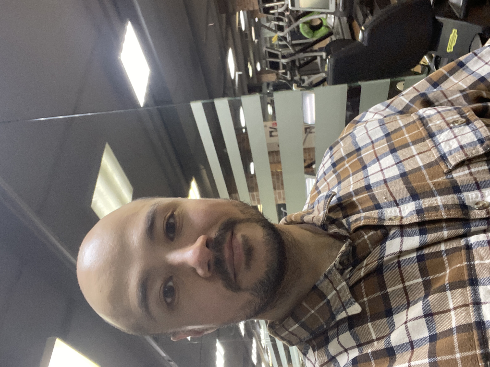 An individual, identified as an adult male, is depicted from the chest up, positioned centrally in the frame, directly facing the viewer. He has a bald head and a neatly trimmed dark beard and mustache. He is wearing a long-sleeved plaid shirt featuring a pattern of brown, white, and dark blue squares. The setting appears to be an indoor fitness facility, specifically a gym.
To the subject's right, a reflective glass or mirror surface reveals a wide array of exercise equipment, including multiple cardio machines such as treadmills and ellipticals, and a black-seated strength training machine. A bright green object is visible further in the background. Overhead, multiple rectangular fluorescent or LED light fixtures illuminate the space. To the subject's left, a glass partition with horizontal opaque stripes partially obstructs the view of the background. The ceiling is dark. The specific location within the city of Izhevsk, Russia, cannot be determined from the visual evidence provided.