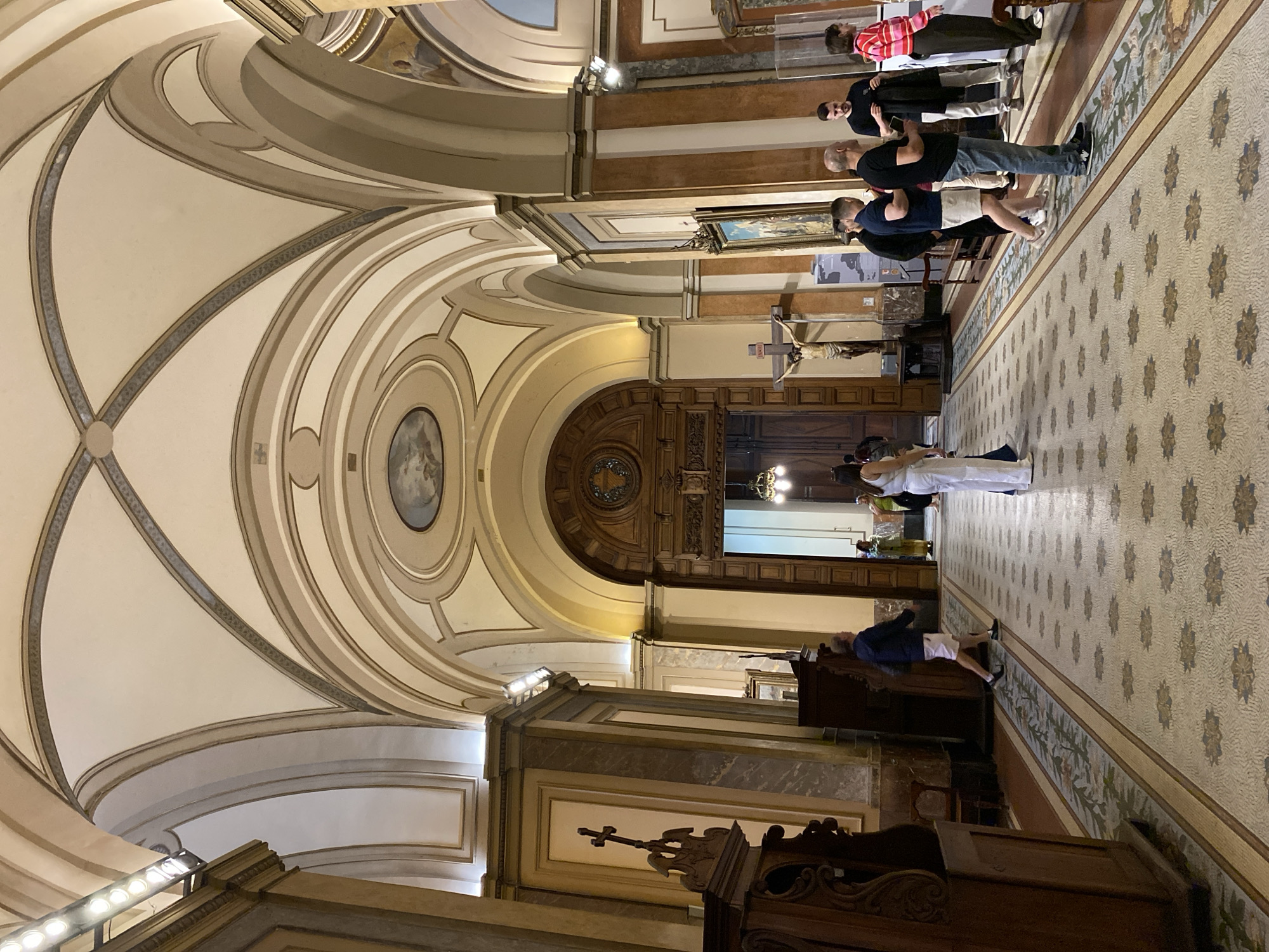 The image captures the interior of a large, ornate church or cathedral, viewed from a side aisle looking towards the main entrance or an altar area. The perspective highlights the impressive vaulted ceiling, adorned with intricate cream and grey patterned moldings and painted details, including a central circular fresco.
The floor is covered with a detailed mosaic tile pattern, featuring alternating sections of geometric designs and floral motifs in shades of brown, beige, blue, and green, running the length of the aisle. Along the right side of the aisle, a group of approximately six adults stands, some observing their surroundings. One person in white attire is visible further down this aisle. On the left side, another individual in a dark blue shirt and white shorts stands near a substantial dark wooden structure, possibly a pulpit or part of the choir stalls, which features carved details and a small cross.
Religious elements are prominent, including a large crucifix mounted on a wall and several framed paintings hanging above marble and wood wainscoting. The far end of the church features grand, intricately carved wooden doors, suggesting the main entrance or access to another important section. Artificial lighting from sconces and unseen sources illuminates the interior, highlighting the architectural details and creating a serene atmosphere. The individuals appear to be visitors or attendees, calmly observing the surroundings. No discernible text is visible in the image.