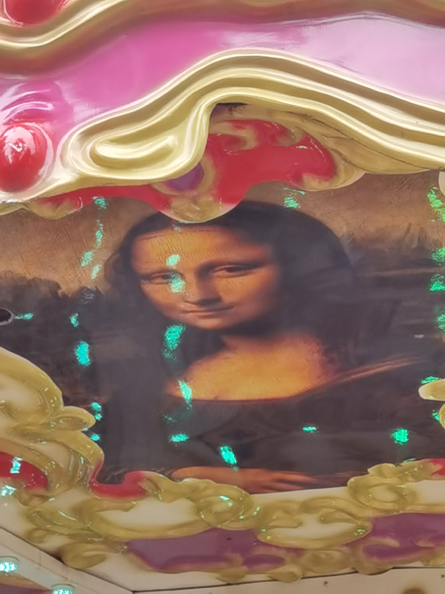 The image shows a reproduction of the Mona Lisa painting, which is framed by a colorful, decorative element that appears to be part of a carousel or a fairground attraction. The framing features swirling patterns in pink, gold, and red, with some areas reflecting light. The Mona Lisa itself is somewhat obscured by scattered green lights, giving it an ethereal or somewhat distorted appearance. The background of the painting is a muted, earthy tone. The lower portion of the image displays more of the decorative framing, which is also brightly colored with purple and gold accents. There are no people visible in the image, and the scene is static, depicting a piece of artwork integrated into a carnival-like structure. The location is specified as Chaikovsky, Russia, suggesting this attraction is in that city.