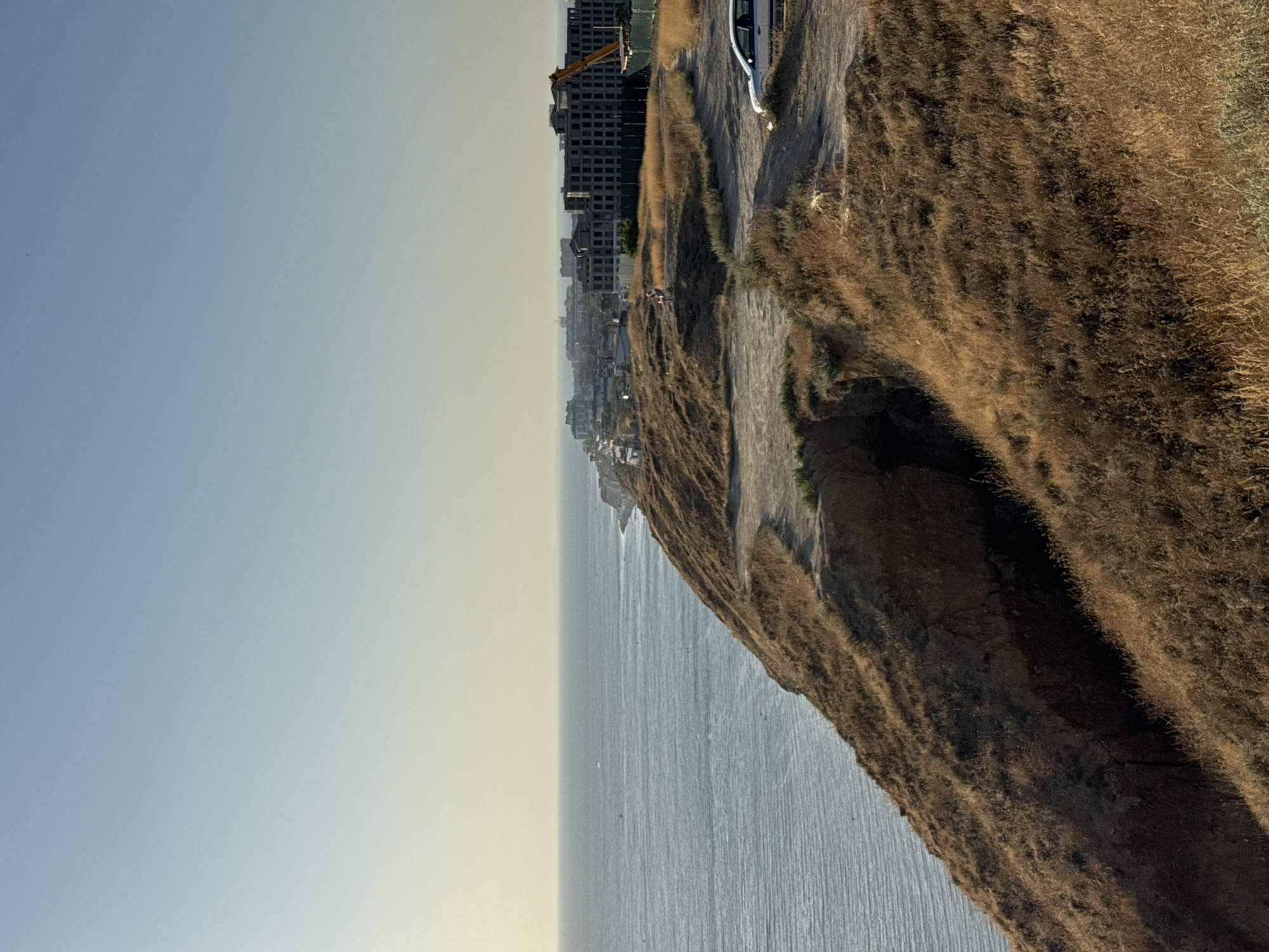 The image depicts a coastal cliffside with a view of the sea and a distant cityscape. The cliff is covered in dry, golden grass and shows signs of erosion, with a deep gully visible. A dirt path winds along the edge of the cliff.
In the middle ground, several buildings are under construction, including a large, unfinished multi-story structure and a yellow construction crane. A few people are visible near the construction site, and a white car is parked on a dirt road in the foreground. The sea stretches to the horizon, with subtle ripples on its surface. The sky is a clear, pale blue, transitioning to a lighter shade near the horizon.
This scene is located in Anapa, Russia, specifically on the coastal cliffs overlooking the Black Sea. The construction activity suggests ongoing development in the area. The natural environment, characterized by the arid cliff vegetation and the expansive sea, forms a stark contrast with the man-made structures.