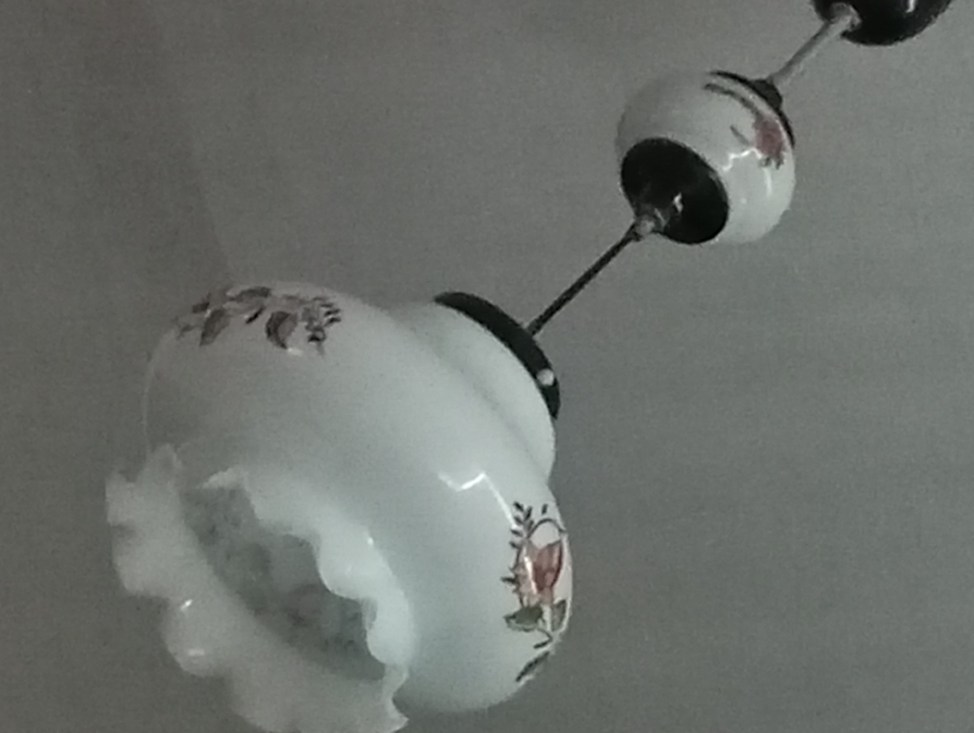 The image shows a white pendant light fixture hanging from a ceiling. The light fixture has a ruffled edge at the bottom and is decorated with floral patterns. There is another, smaller bulb-shaped light fixture above it, also white with floral accents. The background is a plain, light gray wall or ceiling. The lighting is dim, and the image has a soft focus. There are no people or animals present in the image. The location in the city of Soroca, Moldova is not discernible from the image.