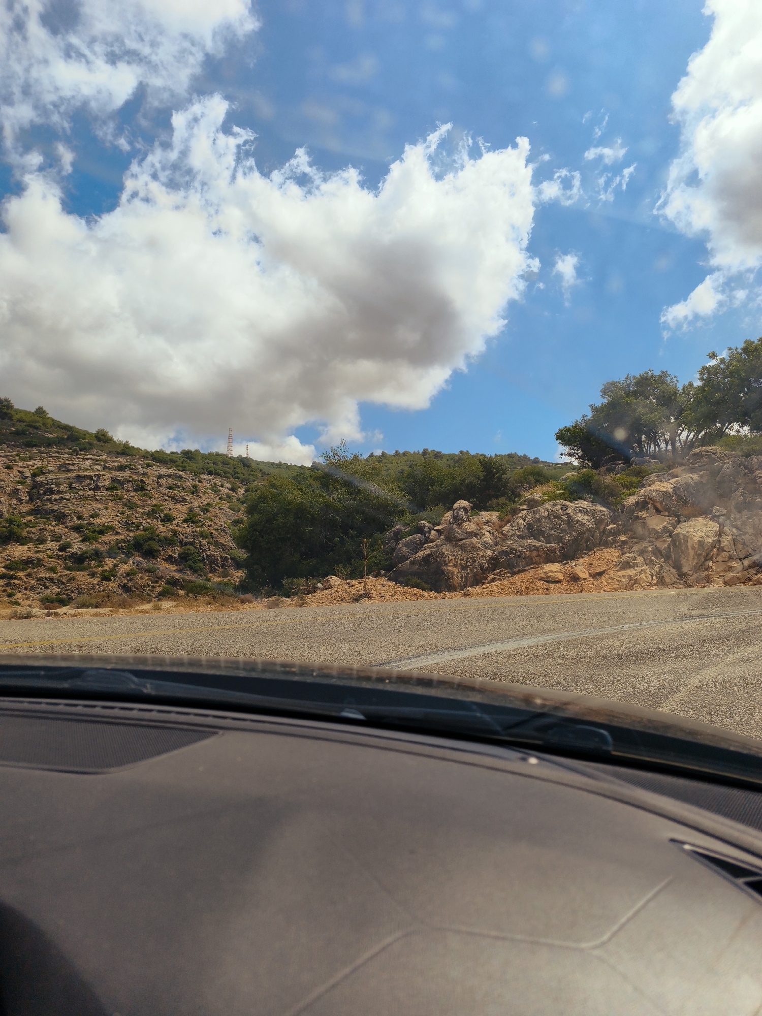 A wide-angle shot captures a view from inside a vehicle driving on a winding road in the Mate Asher Regional Council, Israel. The road, paved with asphalt, curves to the right, with white dashed lines marking its lanes. Beyond the road, the landscape is rugged and mountainous, characterized by rocky terrain and sparse vegetation, with some bushes and small trees scattered across the slopes. Further up the hillside, a cluster of larger trees is visible on the right. The sky above is a vibrant blue, dotted with fluffy white clouds. In the distance, on the crest of the hill, two communication towers are faintly visible. The foreground of the image shows the interior of the car, with the dashboard and windshield visible. The lighting suggests it is daytime, possibly late morning or early afternoon, with sunlight illuminating the scene.