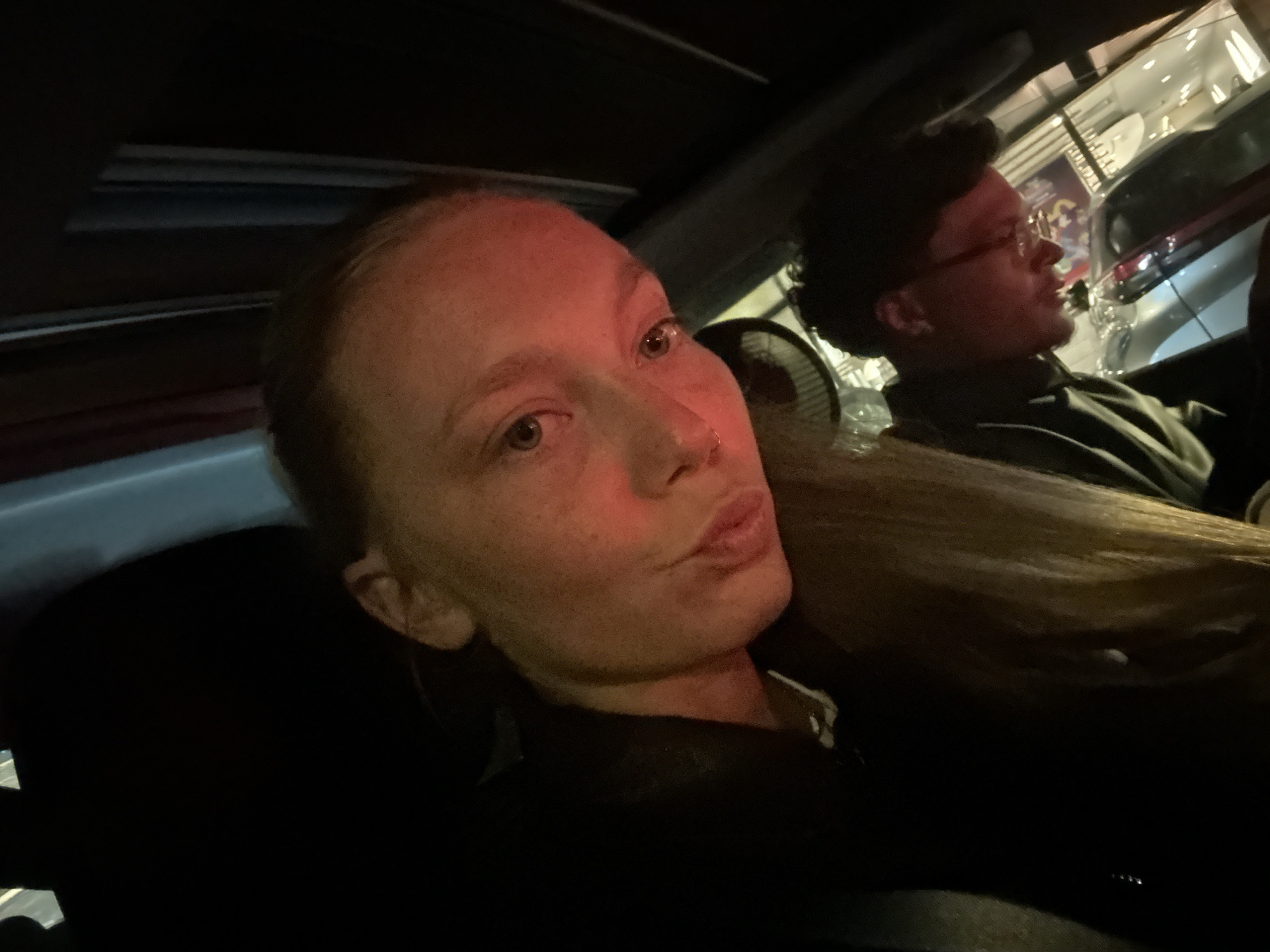 A selfie captured inside a car features two individuals. The foreground is dominated by a woman with light skin and freckles, gazing directly at the camera with pursed lips. Behind her, a man with dark hair and glasses looks off to the side.
Through the car's window, the blurred lights and signs of Noisy-le-Grand, France, are visible, suggesting an urban setting.