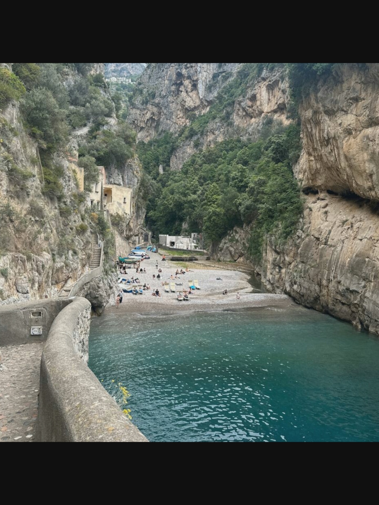 The image depicts a picturesque scene at Fiordo di Furore, a small fjord-like inlet and village on the Amalfi Coast in Italy. A pebble beach lines the turquoise waters of the sea, with several people sunbathing on lounge chairs. Boats are pulled up onto the beach near buildings that cling to the steep, rocky cliffs. A stone walkway curves along the left side of the frame, leading up a set of stairs. The lush green vegetation covers the upper portions of the cliffs, and in the distance, more buildings are visible perched high on the mountainside. The overall impression is one of a secluded and tranquil coastal paradise.