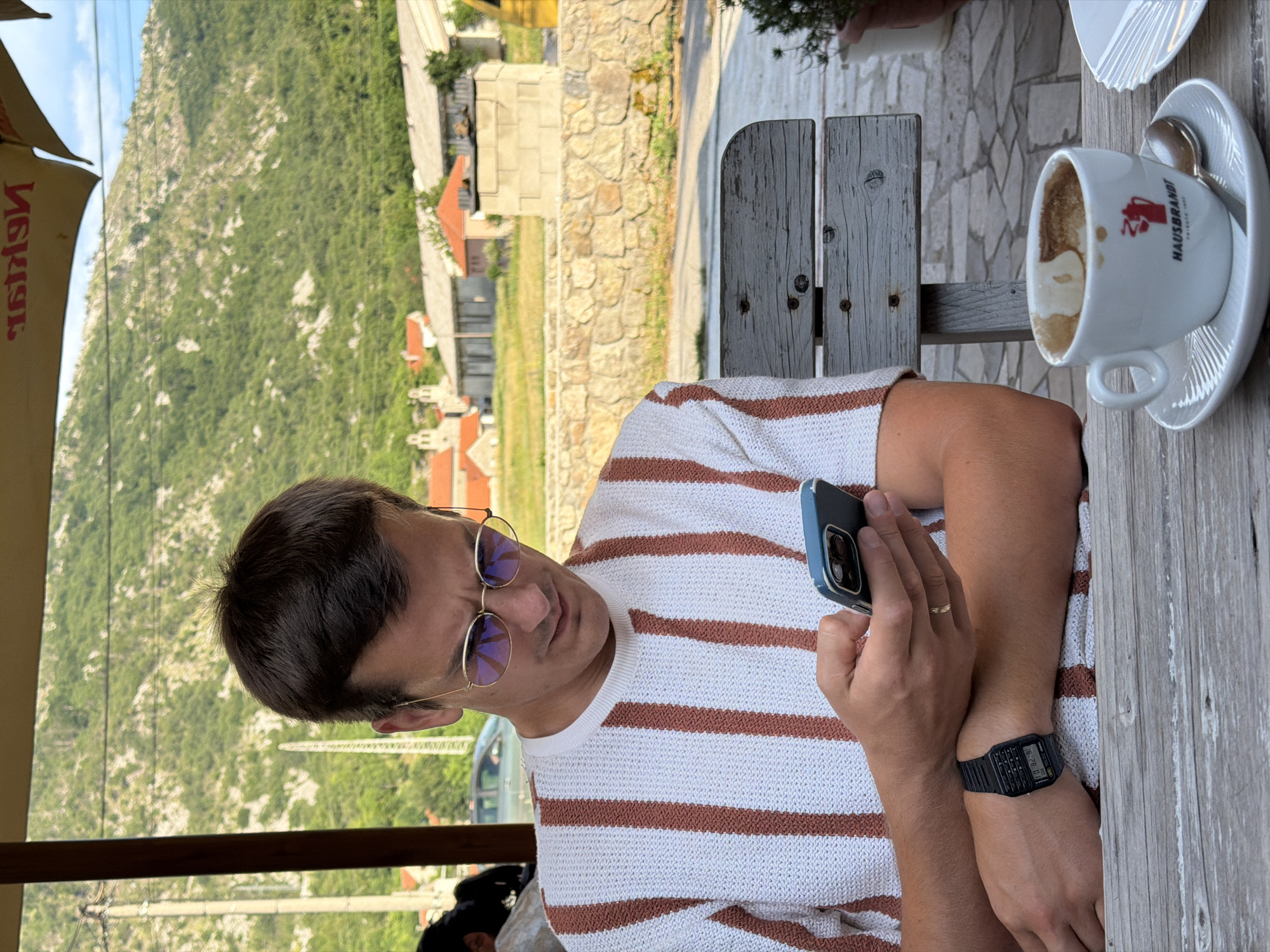 A man is sitting at an outdoor table, looking at his phone. He is wearing a white and brown striped shirt, round sunglasses, and a digital watch. A cup of coffee sits on the table next to him. In the background, a steep, green mountain slope with scattered buildings is visible. The scene appears to be a cafe or restaurant in Njeguši, Montenegro, possibly in the late afternoon given the lighting. The man seems relaxed and engaged with his phone. A logo on the coffee cup is partially visible and says "HAUSBRANDT".