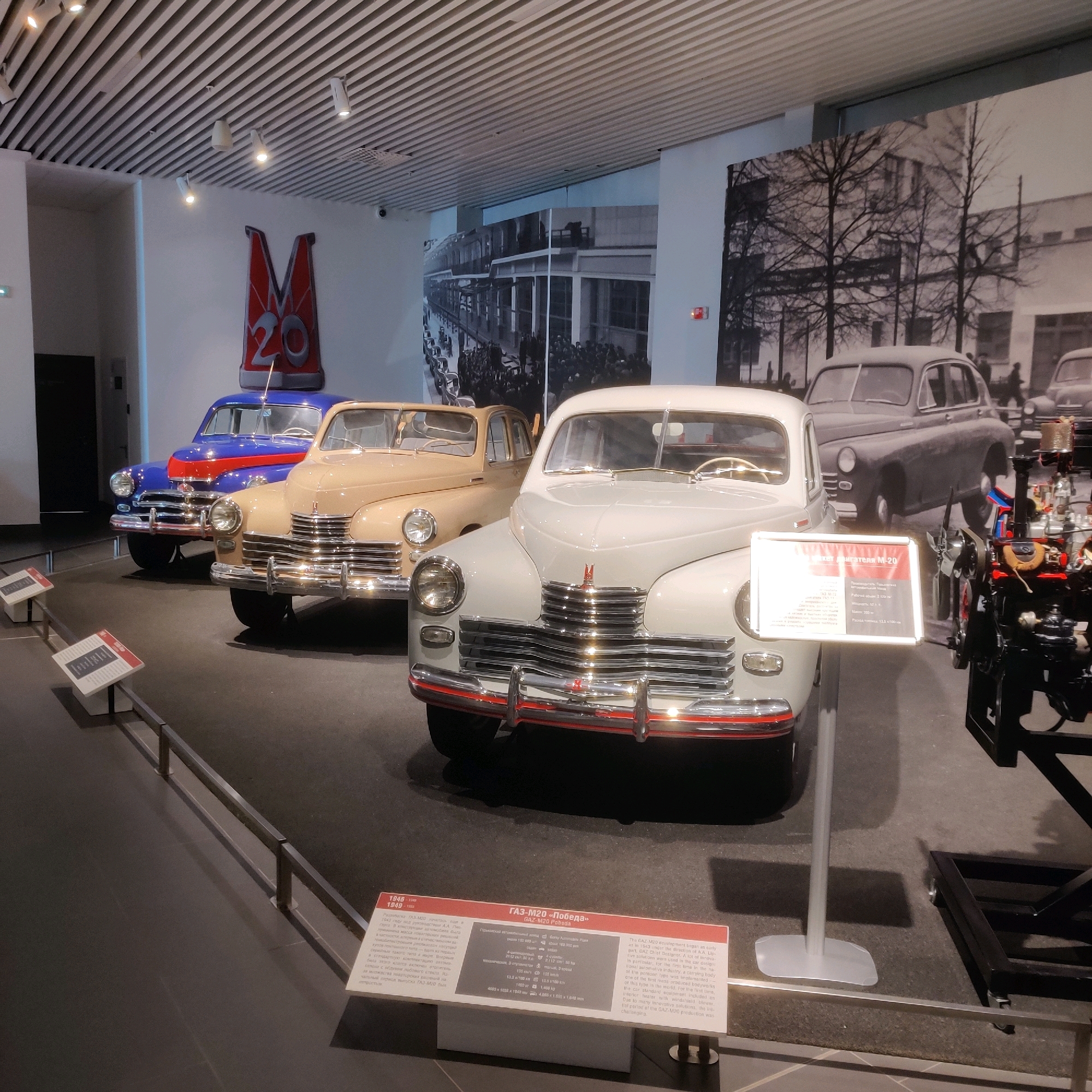 The image shows a museum exhibit featuring several vintage Soviet cars. In the foreground, a light-colored car, possibly a GAZ-M20 Pobeda, is prominently displayed. To its left, a beige car and a blue and red car are parked behind it. A black and white mural depicting a street scene with cars and people is visible on the wall behind the vehicles. The exhibit appears to be well-lit with overhead lighting fixtures. A sign with text and specifications about the GAZ-M20 Pobeda is placed in front of the light-colored car. The overall environment suggests an indoor exhibition space, likely a museum dedicated to automotive history. The museum is located in Verkhnyaya Pyshma, Russia. There are no people actively engaged in the scene, but the exhibit is set up to be viewed by visitors.