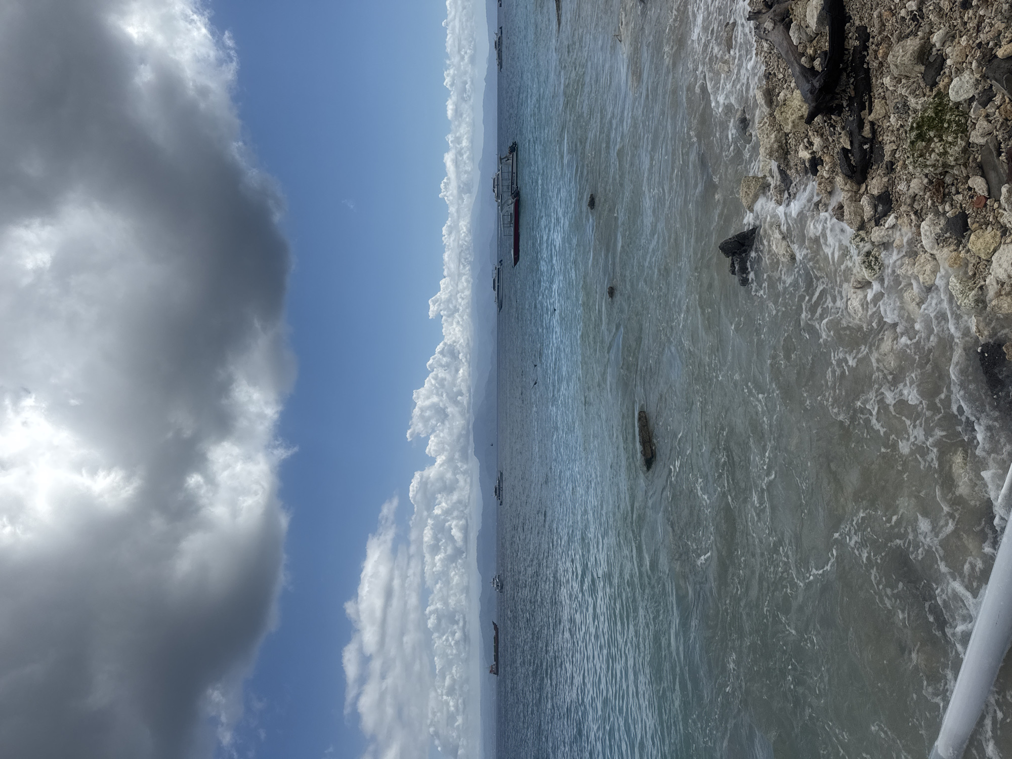 The image shows a shoreline with boats anchored in the distance off Nusa Penida, Indonesia. To the left of the frame, dark, billowing clouds partially obscure a blue sky. The water transitions from a calm, clear blue further out to a foamy, turbulent grey closer to the shore. The shoreline consists of light-colored rocks, some with dark wood debris interspersed. A light-colored pole is visible in the very bottom right corner of the frame.