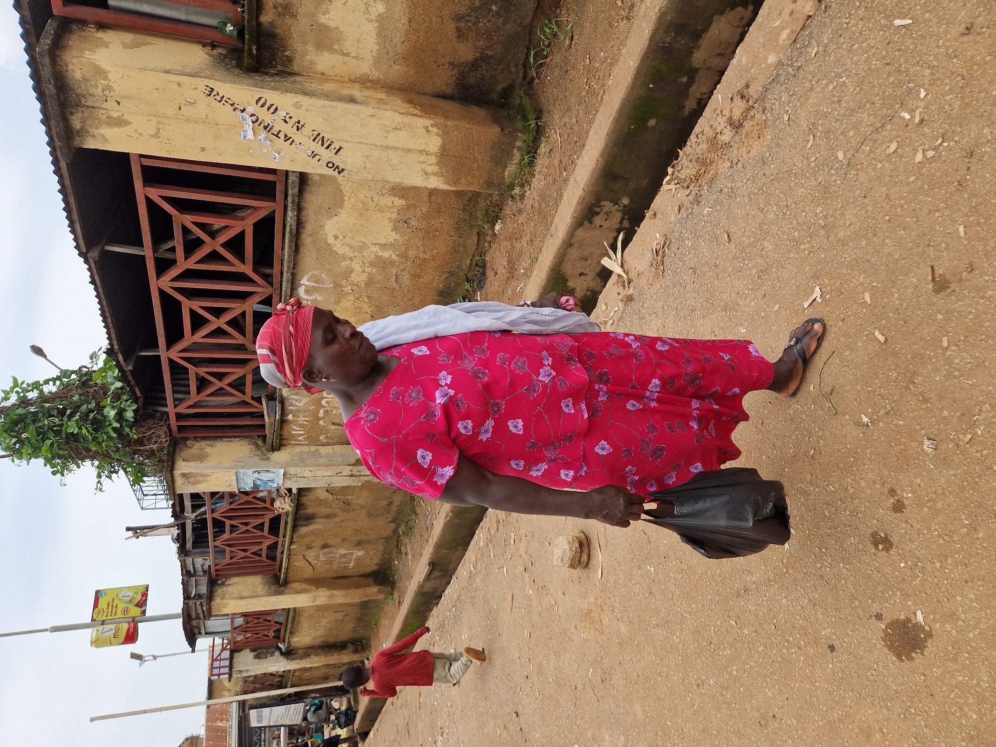 A woman in a red floral dress and headscarf walks on a dirt road in New Stadium, Nigeria, carrying a black plastic bag in her left hand. To her left, a young boy in a red shirt and light-colored pants runs away from the camera. Behind the woman is a weathered, yellowish concrete wall with a wooden lattice fence and a corrugated metal roof structure above it. Graffiti is visible on the wall, including the words "NO URINATING" and "FINE N5,000". The road is uneven and appears to be lined with a drainage ditch on the right side of the image, filled with some debris. The sky is overcast.