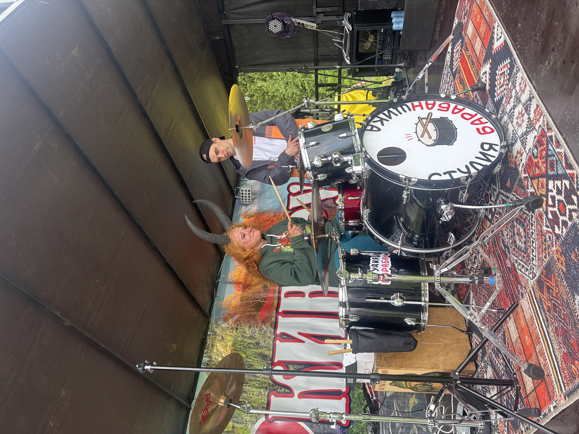 The image depicts an outdoor stage setup, identified by a backdrop banner as located in Большая Кивара, Russia. A female individual with long, bright orange hair and two grey horn-like prosthetics is seated at a black drum kit, actively playing with drumsticks. She is wearing a dark green hooded sweatshirt with a red graphic and teal pants. To her right, a male individual wearing a black baseball cap, a grey, white, and orange jacket, and dark trousers stands observing her, with his hands clasped.
The drum kit consists of black drums with chrome hardware and multiple cymbals, including one marked "PAISTE." The white bass drum head displays a cartoon drum logo flanked by the Russian text "СТУДИЯ БАРАБАНОВ" (Drum Studio) in black and red lettering. The stage floor is dark, partially covered by a multicolored, geometric-patterned rug.
The backdrop banner behind the performers features a vibrant autumnal forest scene with large white, red-outlined Russian text reading "БОЛЬШАЯ КИВАРА" (Bolshaya Kivara). To the far left, a light-colored wooden cajon with a sticker depicting a green frog is visible next to a drumstick holder. On the right side of the stage, various audio equipment including speakers, microphone stands, coiled cables, and a rack unit are present. The stage is covered by a dark, possibly canvas, overhead canopy.