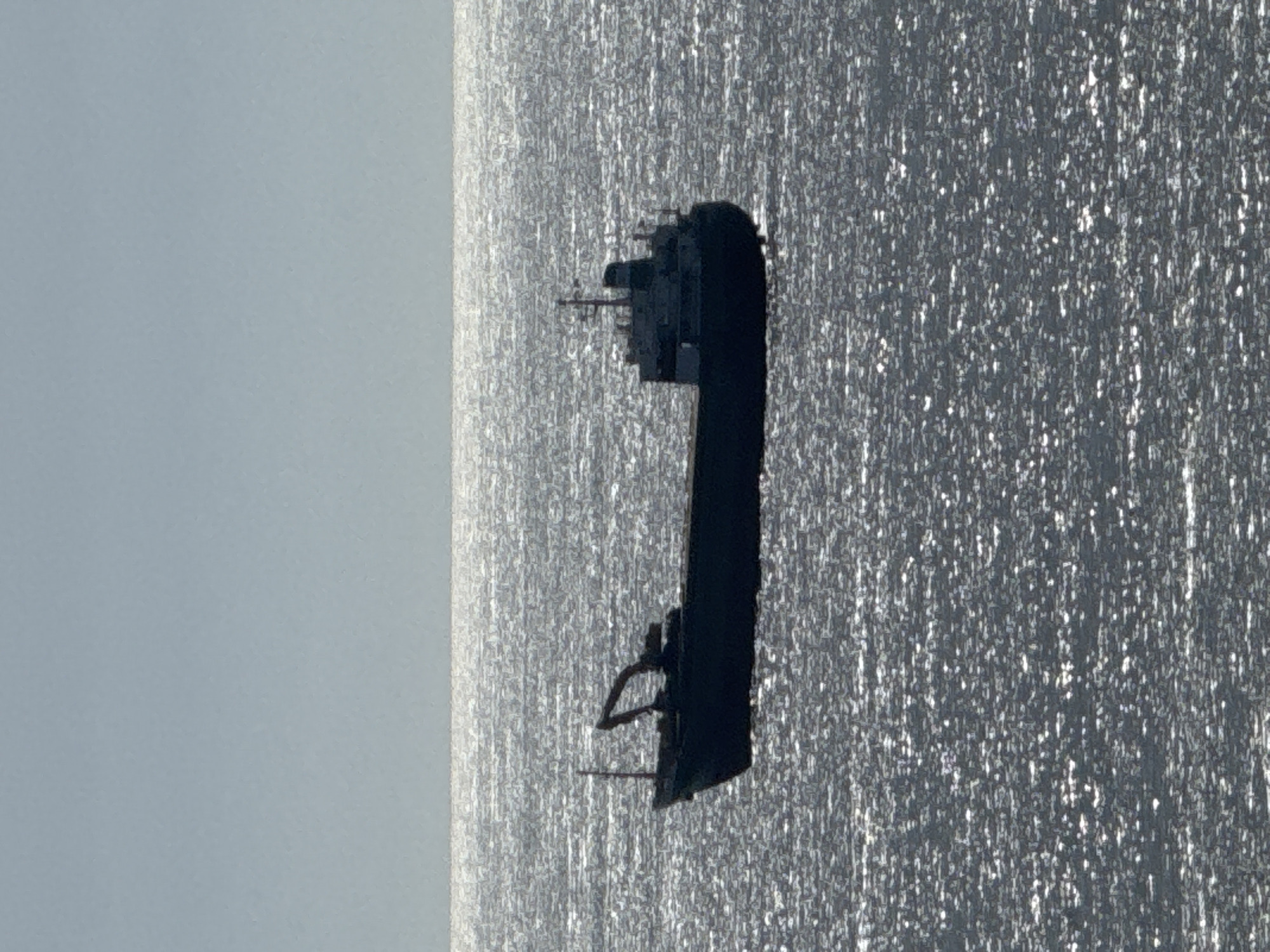 The image, when viewed in its intended horizontal orientation, depicts a large vessel, likely a cargo ship or tanker, silhouetted against a shimmering sea and a pale sky. The ship is positioned horizontally across the mid-ground, appearing dark in color. Its superstructure, featuring multiple antenna-like elements and intricate details, is visible towards the stern end (right side of the ship's length). Some railing or structural components can also be observed at the bow (left side of the ship's length).
The water surface beneath the ship is intensely reflective, covered in countless bright glints of sunlight, suggesting a calm sea under direct, bright daylight conditions, possibly midday or early afternoon. The sky above is a uniform light blue-grey, hinting at clear or thinly overcast weather.
No specific activity or individuals are discernible on board the ship. There is no visible text anywhere in the frame. While the location context is provided as Varna, Bulgaria, there are no specific visual cues within the image itself to confirm this geographical detail.