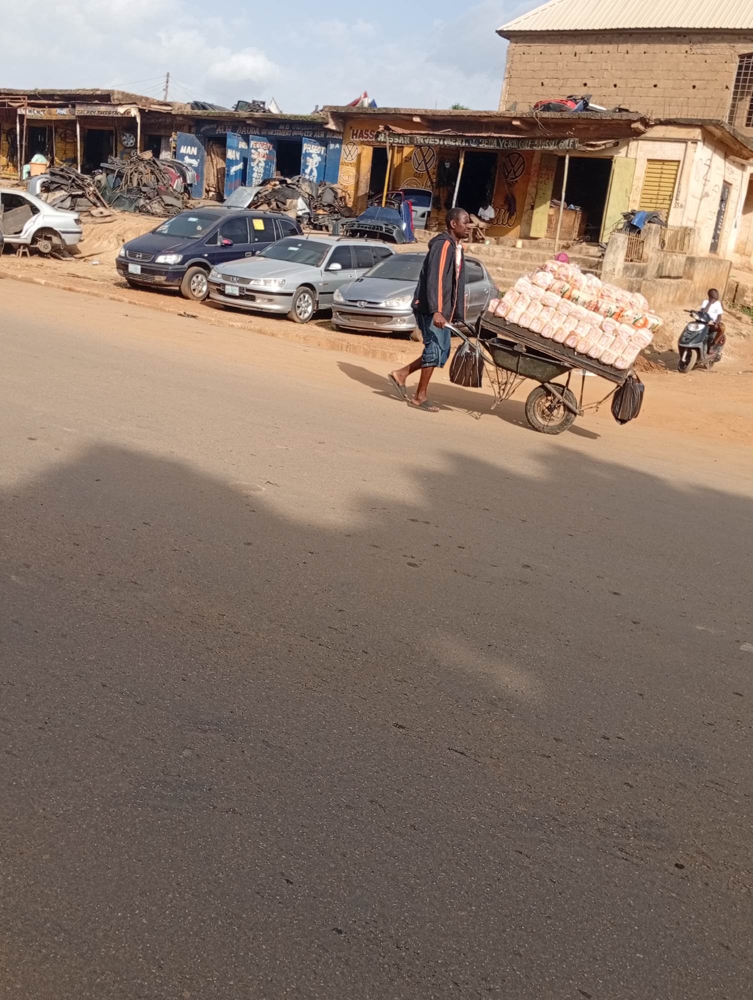 This is a photograph taken in Rafin Par, Nigeria, depicting a street scene with a man pushing a cart loaded with goods. The man, wearing a black jacket with orange trim and denim shorts, is walking on the paved road. The cart he is pushing is a two-wheeled trolley piled high with what appear to be packages of bread. Two black bags are attached to the sides of the cart.
Behind the man and the cart, several cars are parked. These include a dark blue minivan, a silver sedan, and another silver car. Further back, a row of market stalls or shops lines the street. These structures appear to be made of brick and corrugated metal. Signs with various text, some in English and some in other languages, are visible above the shops, suggesting they sell car parts and other merchandise. One sign is particularly legible and reads "MAN" in large blue letters.
To the right of the frame, a young person on a motor scooter is visible, riding away from the camera. The overall environment suggests an active marketplace or commercial area within a Nigerian town. The ground is dusty, and the lighting indicates it is daytime.