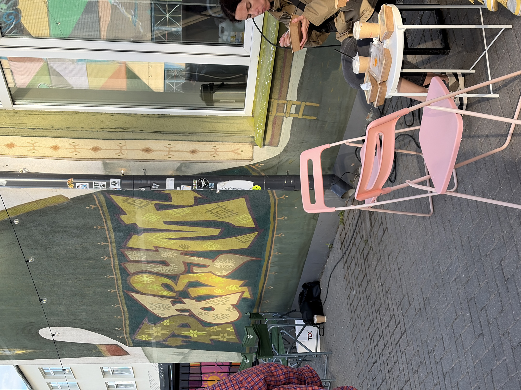 The image shows an outdoor scene in Minsk, Belarus, featuring a person sitting at a small, round, white table with food and drinks. The person is wearing a beige jacket and is seen interacting with the food. There are two pink chairs next to the table. In the background, there's a building with a large, colorful mural that appears to be graffiti-style art in yellow and green shades. A string of lights hangs in front of the mural. On the ground, there's a paved surface, and camping chairs are visible near a white box labeled "TDX". The scene suggests an outdoor cafe or restaurant setting.