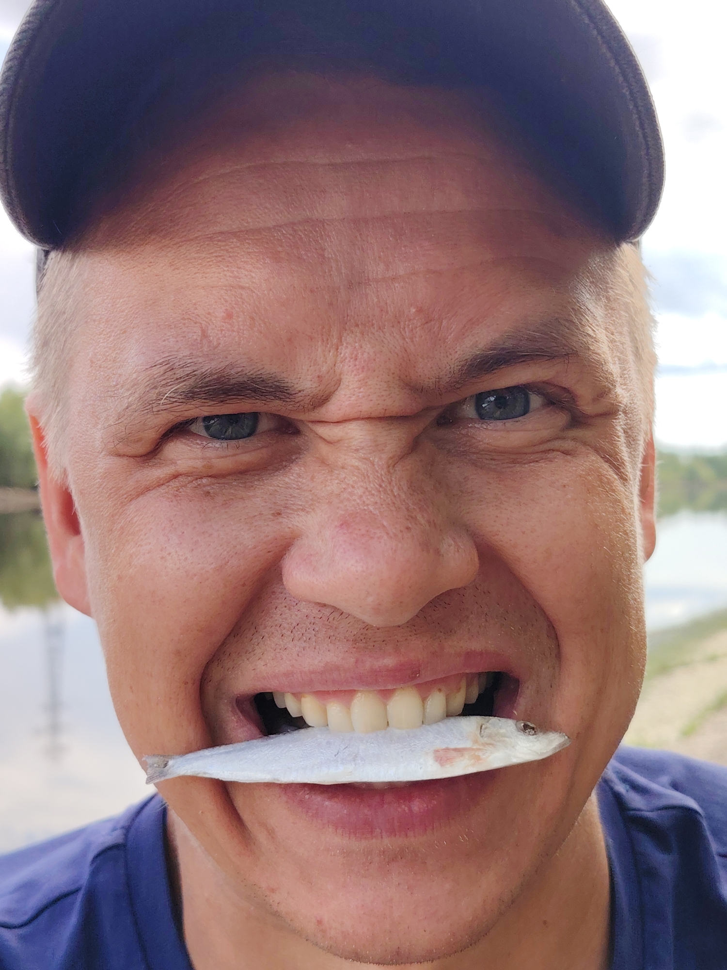 A close-up captures a man with light skin, wearing a blue t-shirt and a dark cap, positioned outdoors near a body of water. He holds a small, silver fish horizontally in his mouth. His face is contorted with a strained expression, furrowed brow, and clenched teeth. The background shows water and some greenery along the shoreline. This image appears to have been taken in the city of Chaikovsky, Russia.