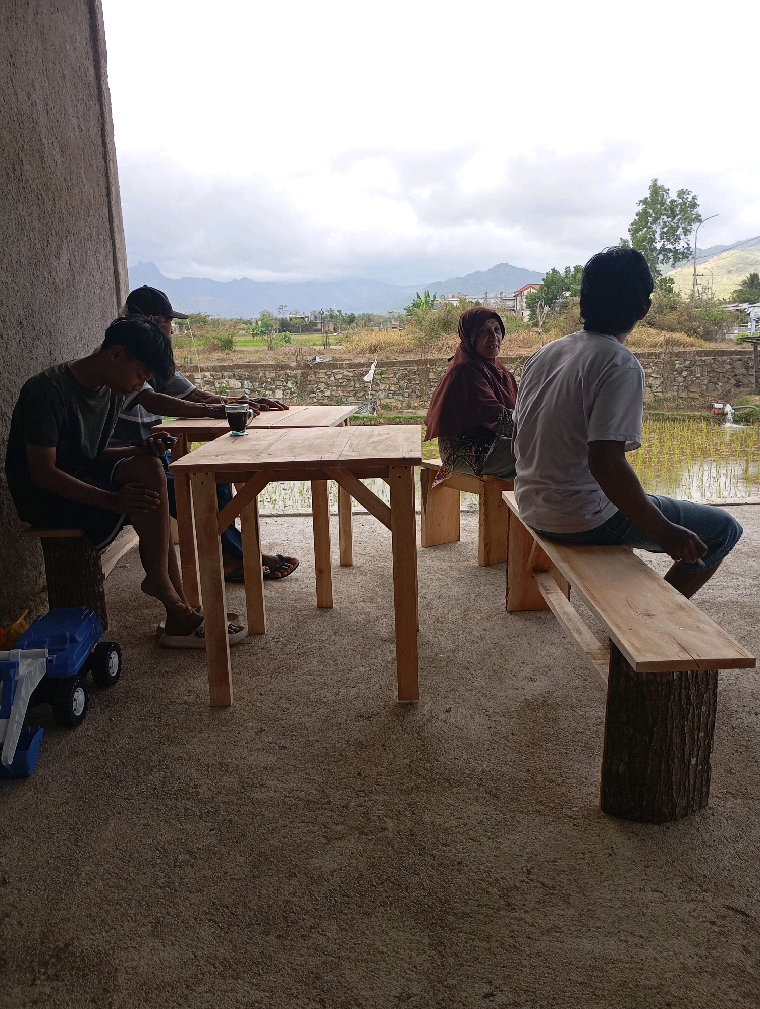 Four individuals are seated at wooden tables and benches outside a building, with a rural landscape and cloudy sky in the background. The setting appears to be a casual outdoor dining or rest area, possibly a cafe or warung, located in the city of Pacitan, Indonesia.
In the foreground, on the left, a young man wearing a dark t-shirt and shorts sits on a wooden bench. He is looking down at his phone, and a blue toy truck is on the ground next to him. Two other men are seated at a wooden table behind him. One is wearing a black baseball cap and a grey t-shirt, and the other is wearing a dark t-shirt. They appear to be engaged in conversation or looking at something on the table. A cup of coffee is visible on the table.
Further back, a woman wearing a maroon headscarf and a patterned dress is seated on a wooden bench at another table. She is looking towards the right. A man in a light grey t-shirt and blue jeans is seated on a long wooden bench to the right, facing away from the camera. He appears to be holding a cigarette.
The environment consists of a concrete floor, simple wooden furniture, and an open view of a rural area. Beyond the seating area, there are rice paddies, stone walls, trees, and distant hills or mountains under a heavily clouded sky. The architecture of the building visible on the left suggests a modest structure with a textured wall. The overall atmosphere is relaxed and informal, indicative of a local establishment in a scenic, rural setting within Pacitan.