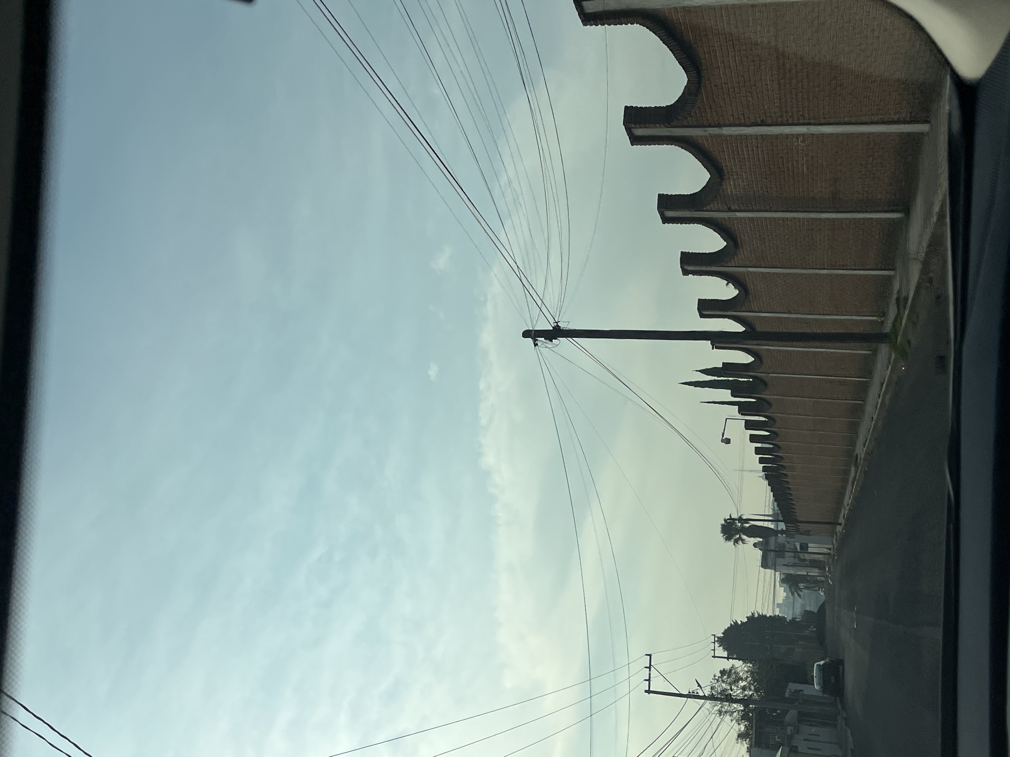 The image depicts a street scene in Puebla, Mexico, viewed from inside a vehicle. A long, brick wall with arched architectural details runs along the right side of the frame. Utility poles and power lines crisscross the sky above the street. The street itself appears to be paved and leads into the distance, where buildings and trees are visible. A car is parked along the right side of the road further down the street. The sky is overcast.