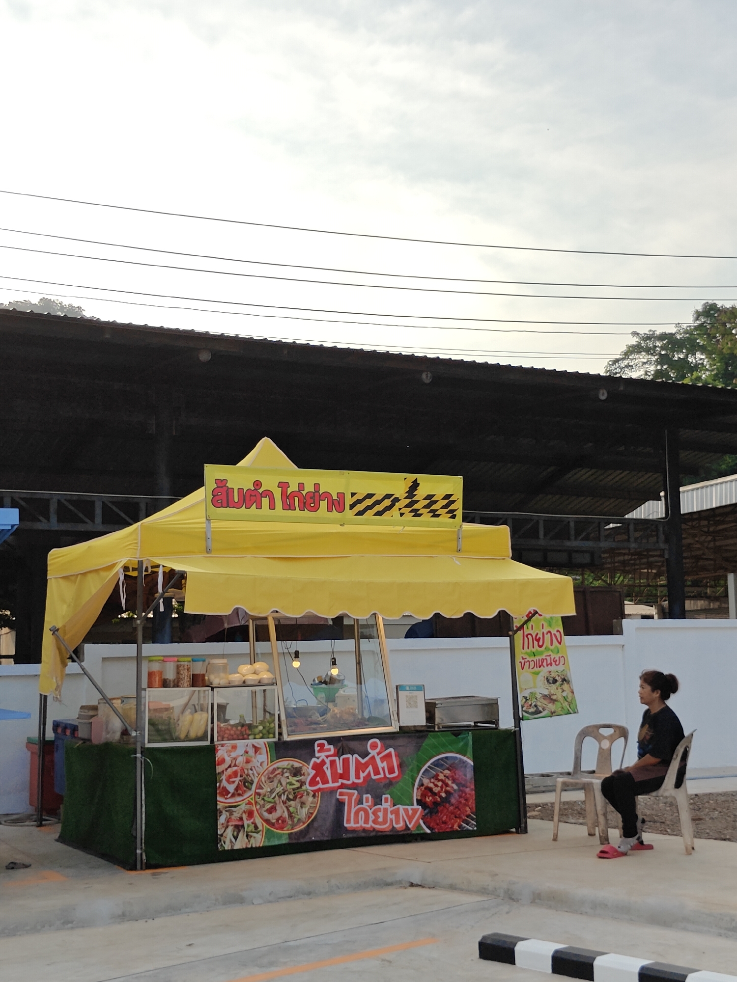 In the city of Mu Si, Thailand, an outdoor food stall is set up under a yellow canopy. The stall displays various food items in glass cases, including what appears to be salads, vegetables, and cooked meats. There are also containers of sauces and condiments. The front of the stall features a large banner with images of food, including a papaya salad and grilled skewers. A woman is seated on a plastic chair to the right of the stall, looking away from the camera. The stall is situated on a paved surface, with a white wall and some greenery visible in the background. Overhead power lines are present in the sky.