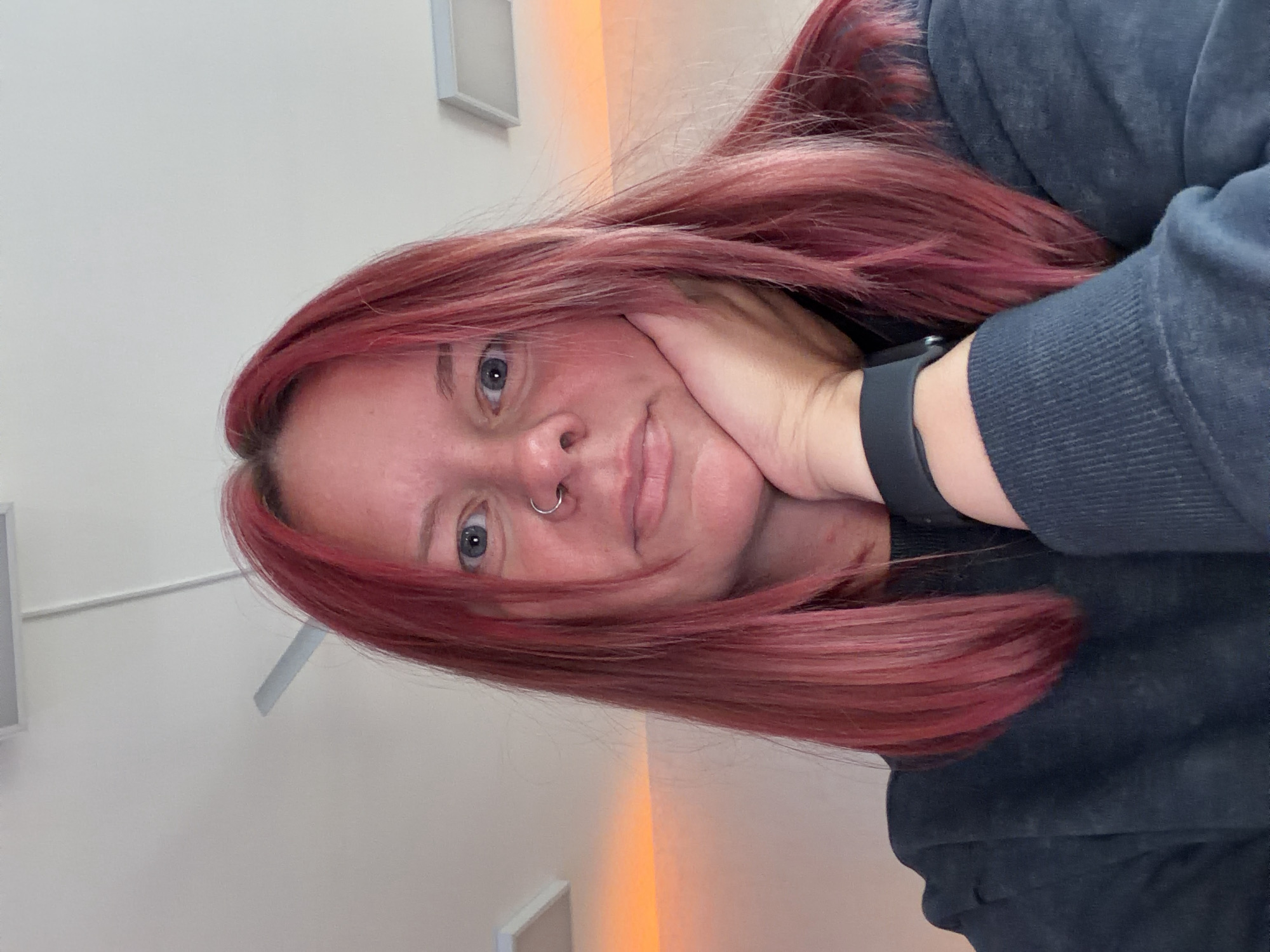 A woman with dyed red hair and blue eyes is shown in a close-up selfie. She has a nose ring and is wearing a black sweatshirt with a smartwatch on her left wrist. Her left hand is supporting her face. The background appears to be an indoor setting with a white wall, a ceiling light fixture, and a vertical orange light strip. There is no information in the image to indicate the location as Chaikovsky, Russia.